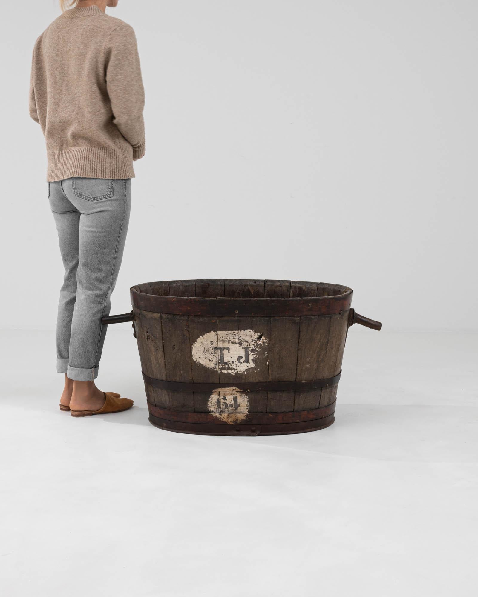 Immerse yourself in the essence of rustic charm with this Early 20th Century French Wooden Grape Container, a genuine piece of agricultural history. Crafted from solid wood, this robust vessel has journeyed through time, its darkened staves bound by