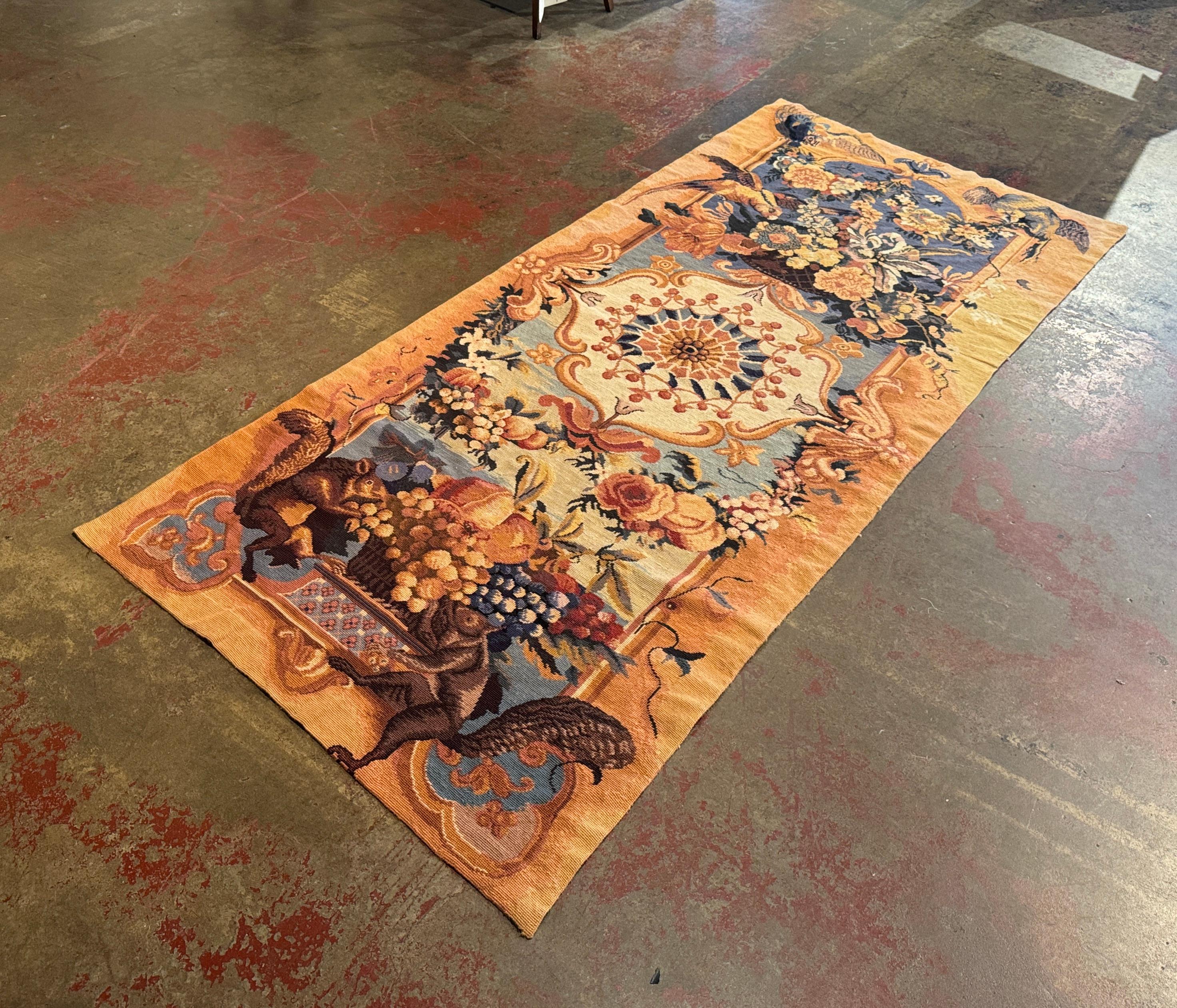 This antique needlepoint tapestry was hand woven in France, circa 1920. Rectangular in shape, the colorful wall decor depicts a large central medallion framed by curling acanthus leaves, floral embellishments, and ornamental flourishes in warm hues