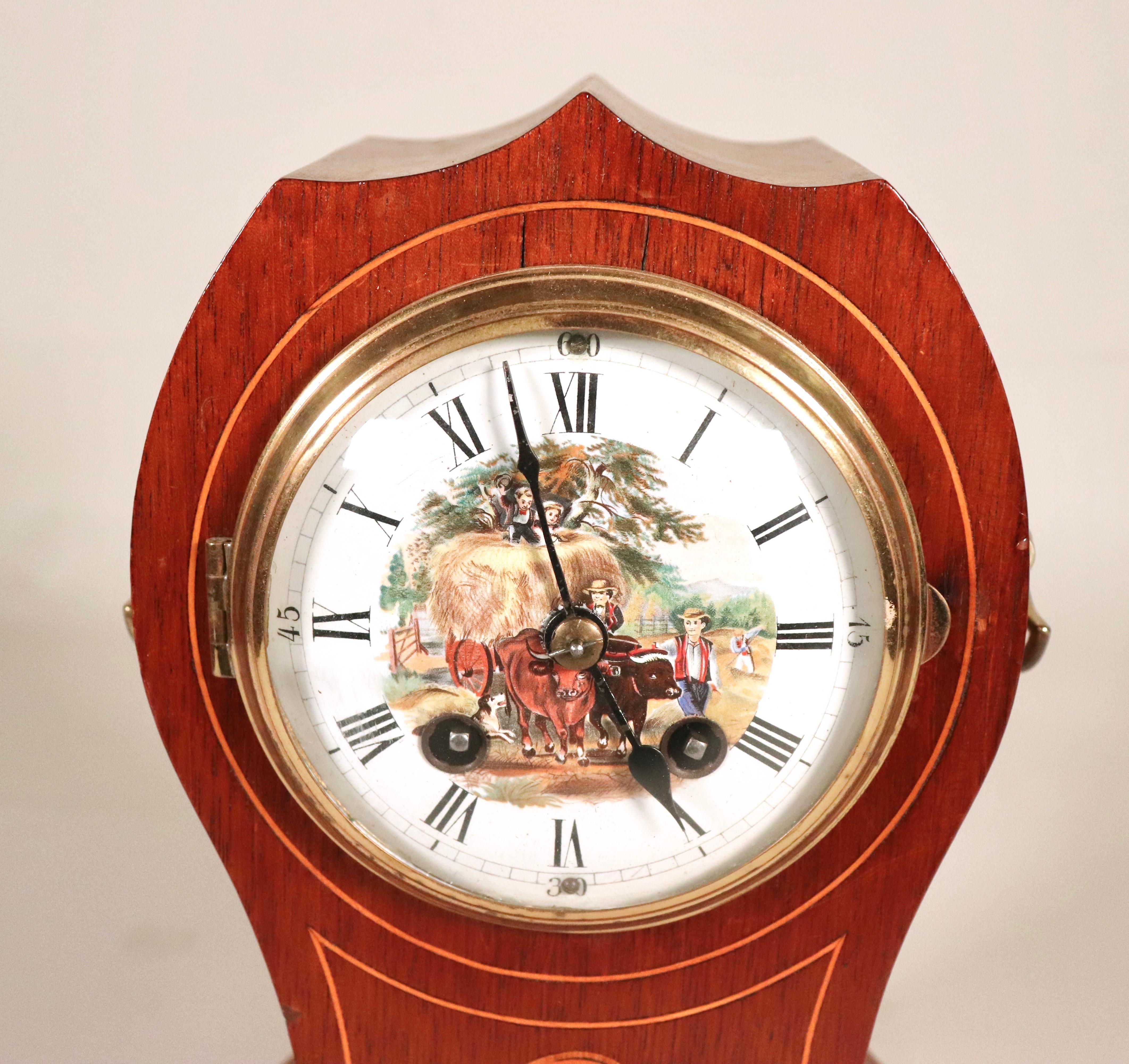 This Early 20th-century German Philipp Haas & Söhne balloon-shaped mantel clock features a mahogany case with boxwood and ebony inlays. The porcelain is hand-painted with a pastoral scene.  The clock is spring-driven and has an eight-day movement