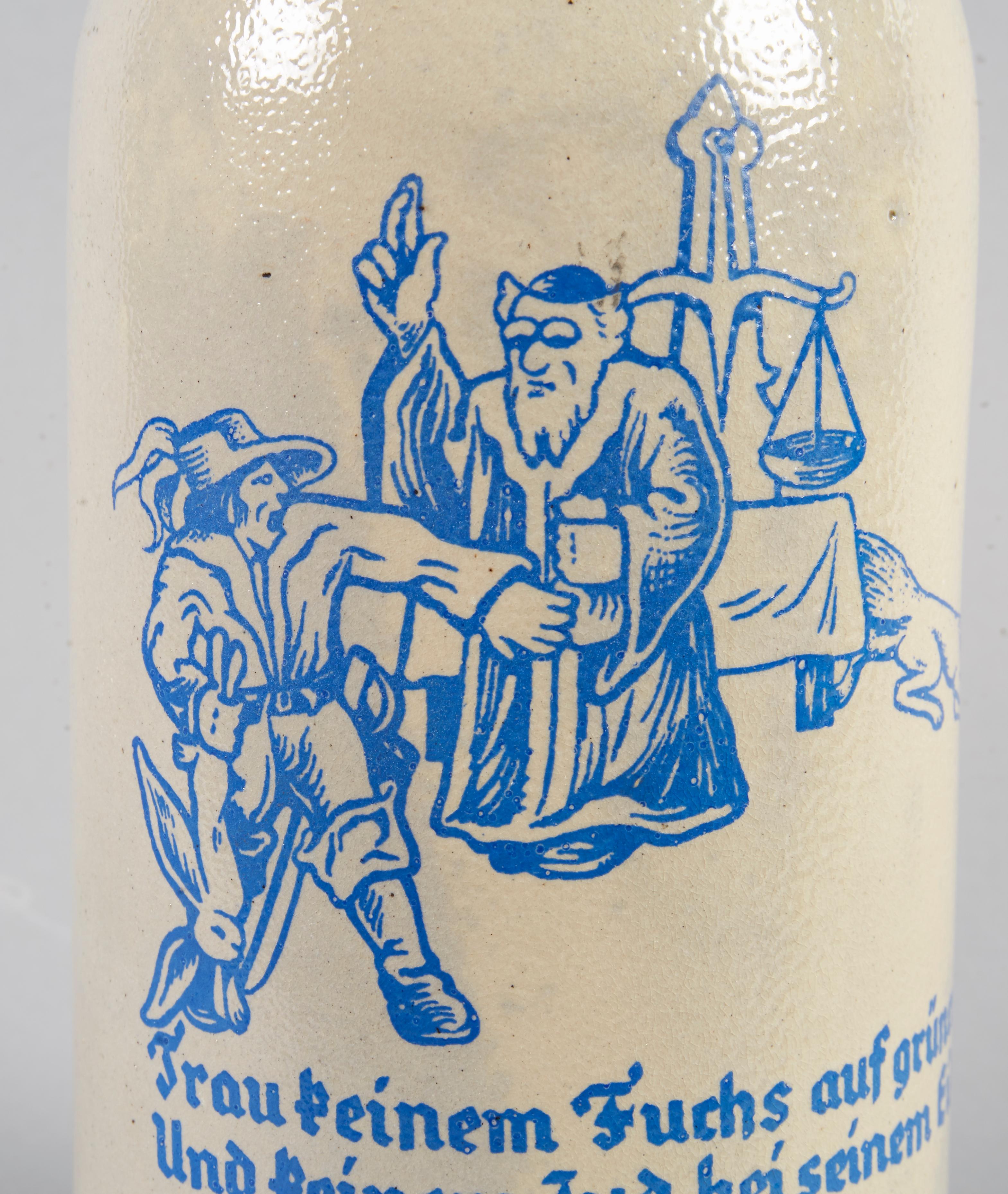 Gray glazed stoneware beer stein with a curved ceramic handle with a domed pewter lid with a pewter thumb-lift. Transfer printed in blue is a scene of a soldier, a religious Jew, scales on a table, and a fox. Titled “Trau keinem Fuchs auf gruner