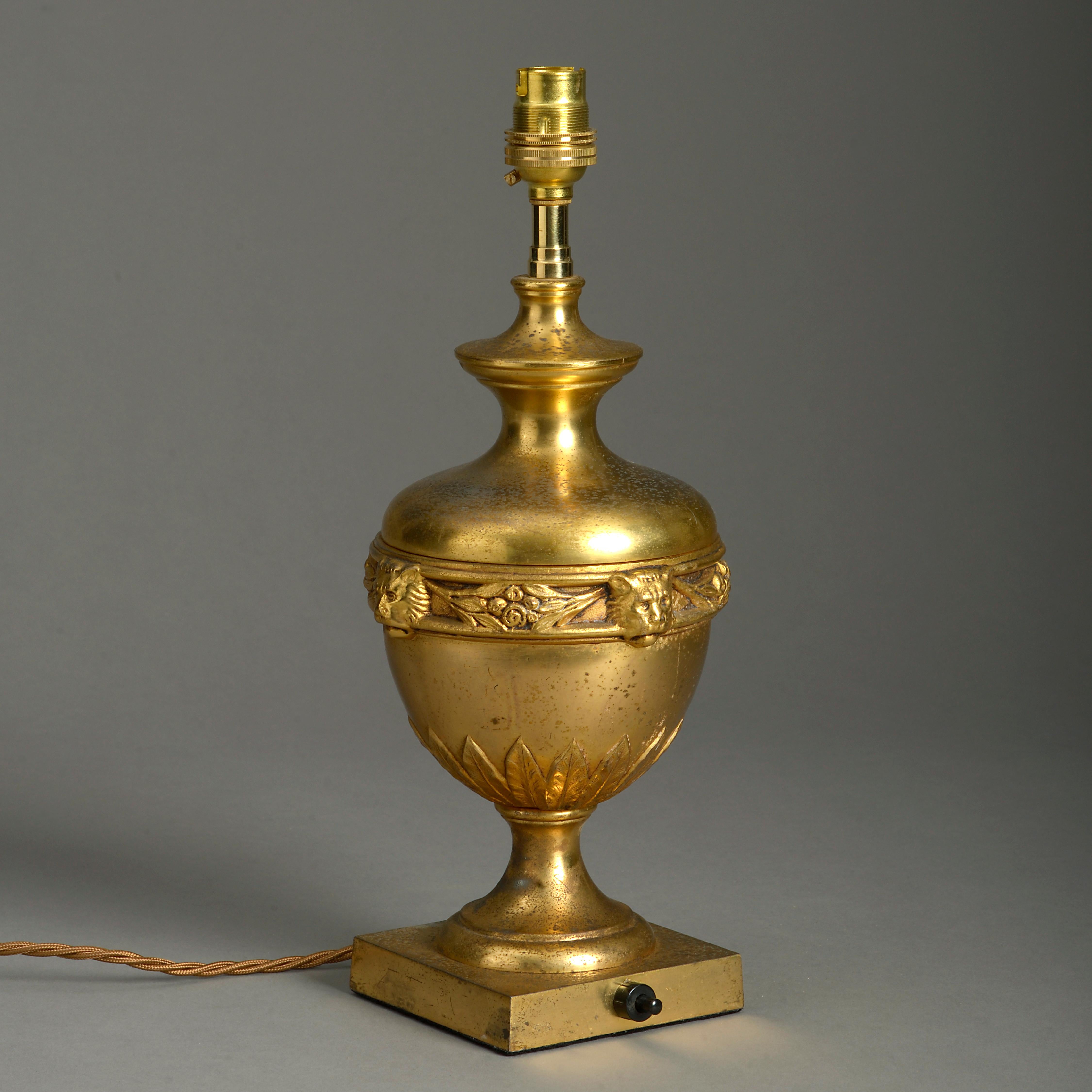 An early 20th century gilt metal urn, having a central band of lion masks articulated with floral swags above palmettes and raised upon a square plinth.

Now wired as a table lamp.

Dimensions refer to vase only and do not include electrical