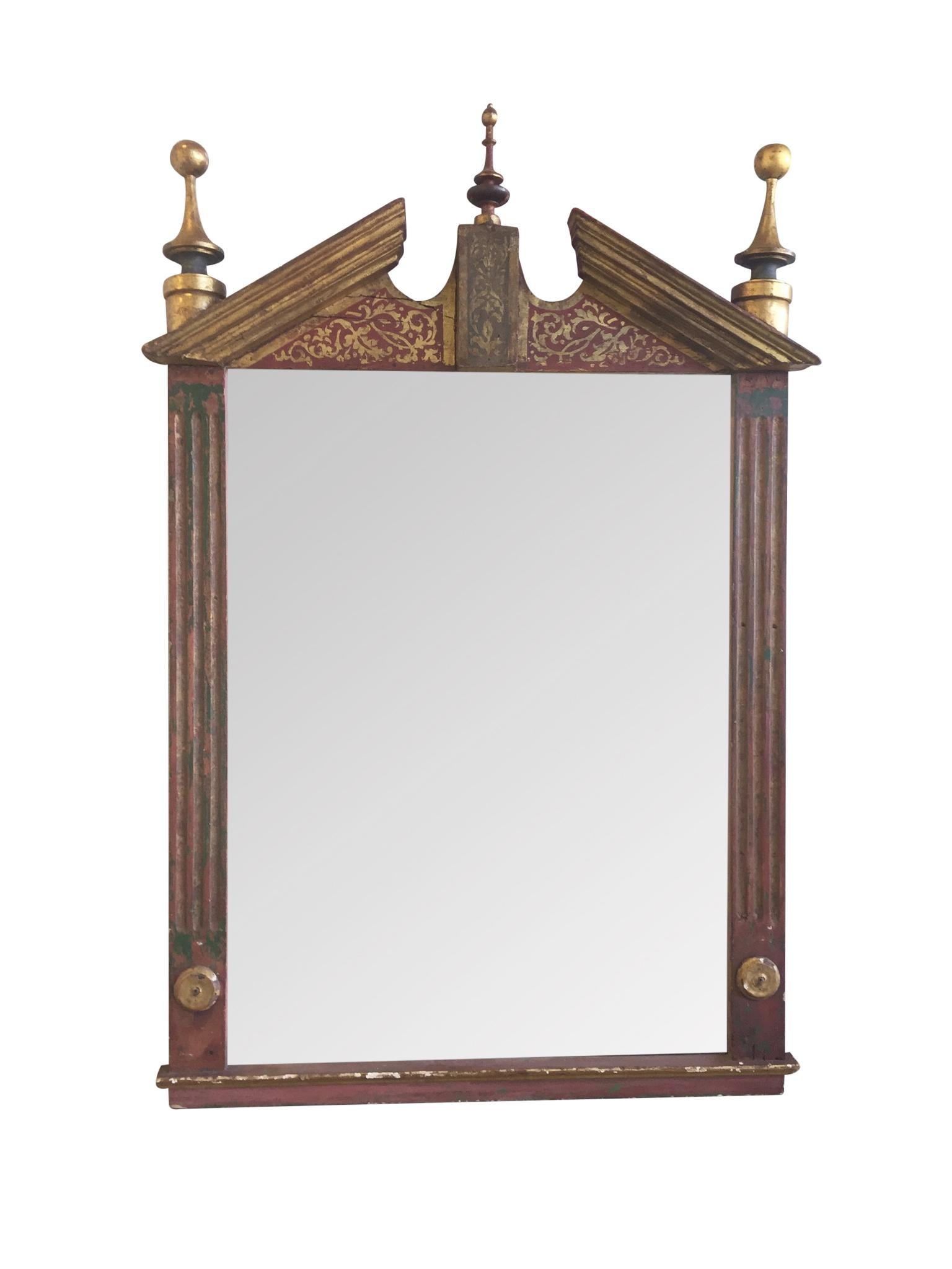 An elegant, finely aged wall mirror in the neoclassical style. Crafted in the early 20th century. The frame is comprised of wood painted with gold and a burgundy hue. The gilt detailing highlights certain elements of the mirror, such as the
