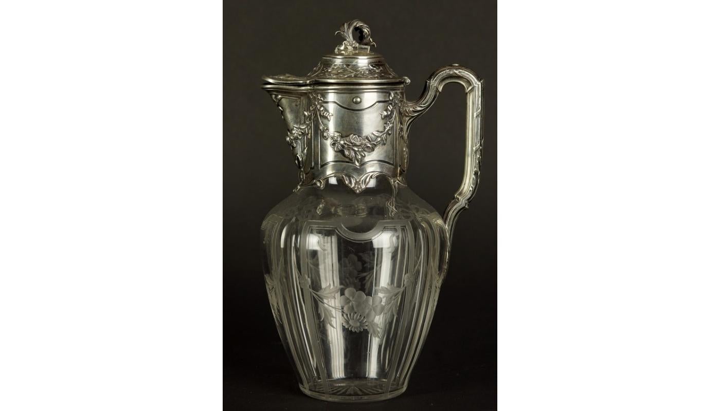 Glass wine jug with silver fittings. Decorating with the use of various plant motifs - acanthus leaves on the handle and pinnacles, laurel twigs on the lid and flower garlands hung on bows in the neck part. The glass belly presents a similar