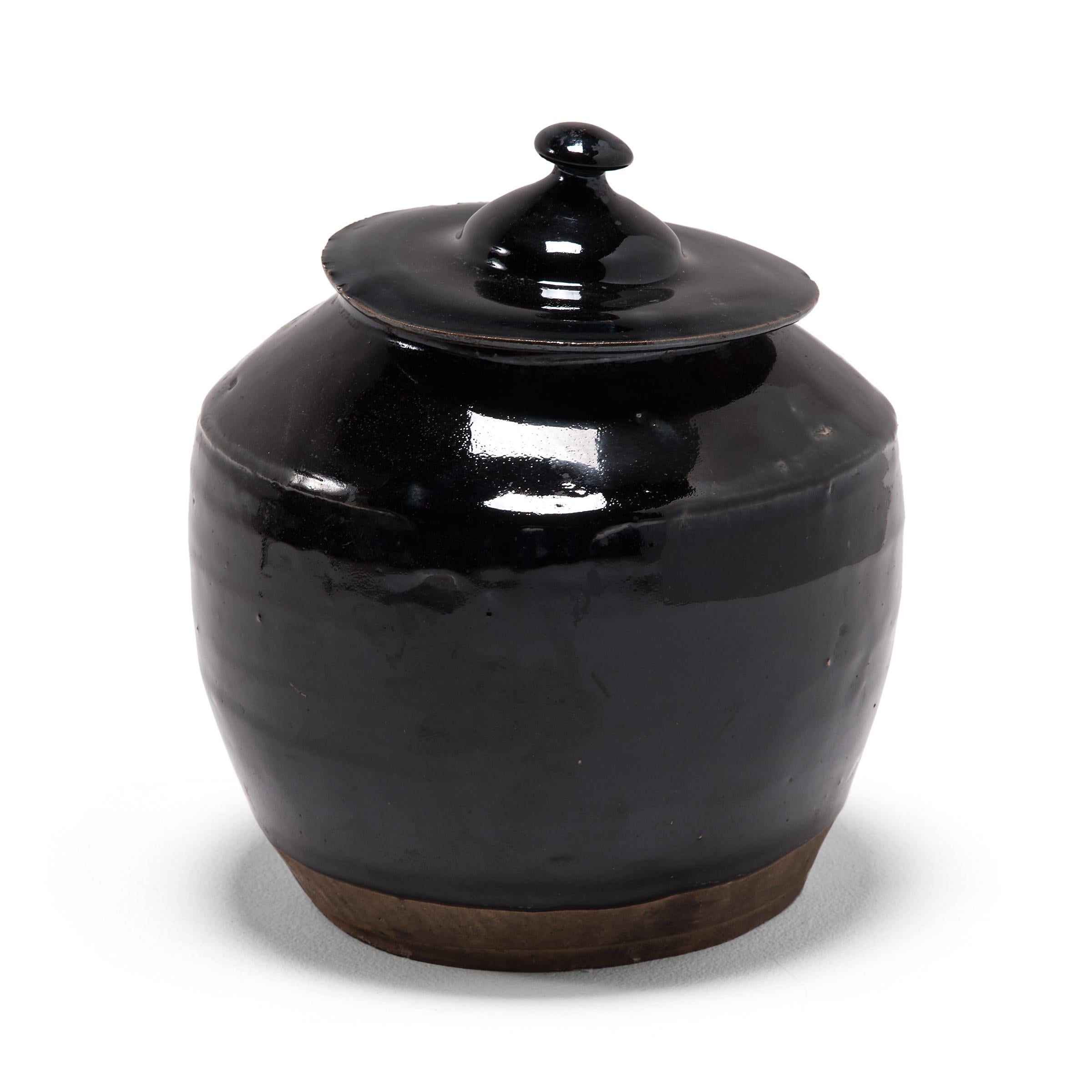 A dark glaze coats the rounded form of this squat kitchen vessel, cloaking the exterior with a glossy shine. The early 20th century jar was once used for storing food in a Qing-dynasty kitchen, as evidenced by its interior glaze. Dotted with