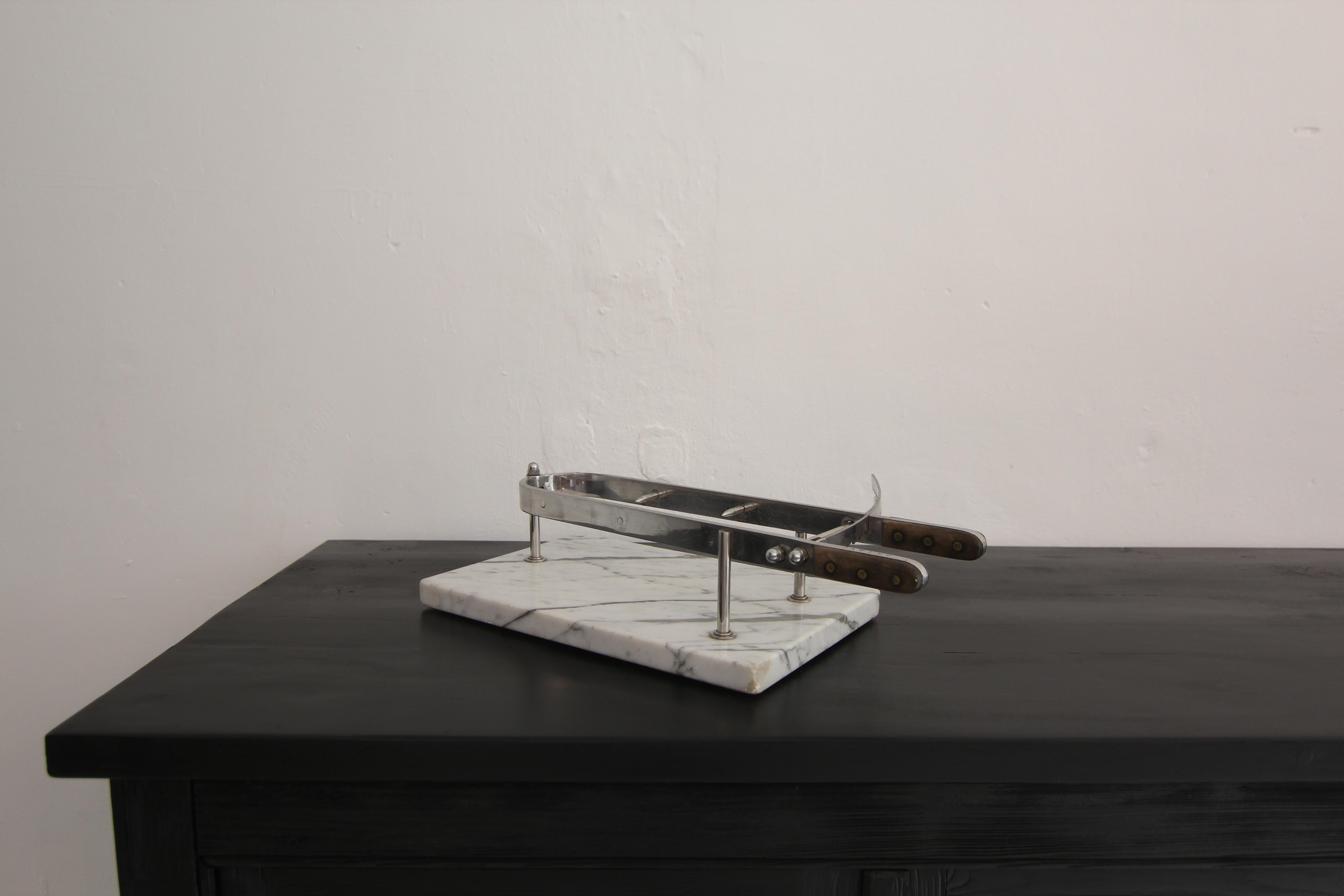 Ham holder made of chrome-plated metal with wood handle grips on white carrara marble base. Probably French or Italian, first half of the 20th century. 
A ham holder is an instrument used to hold hams in place so that they can be cut more easily.