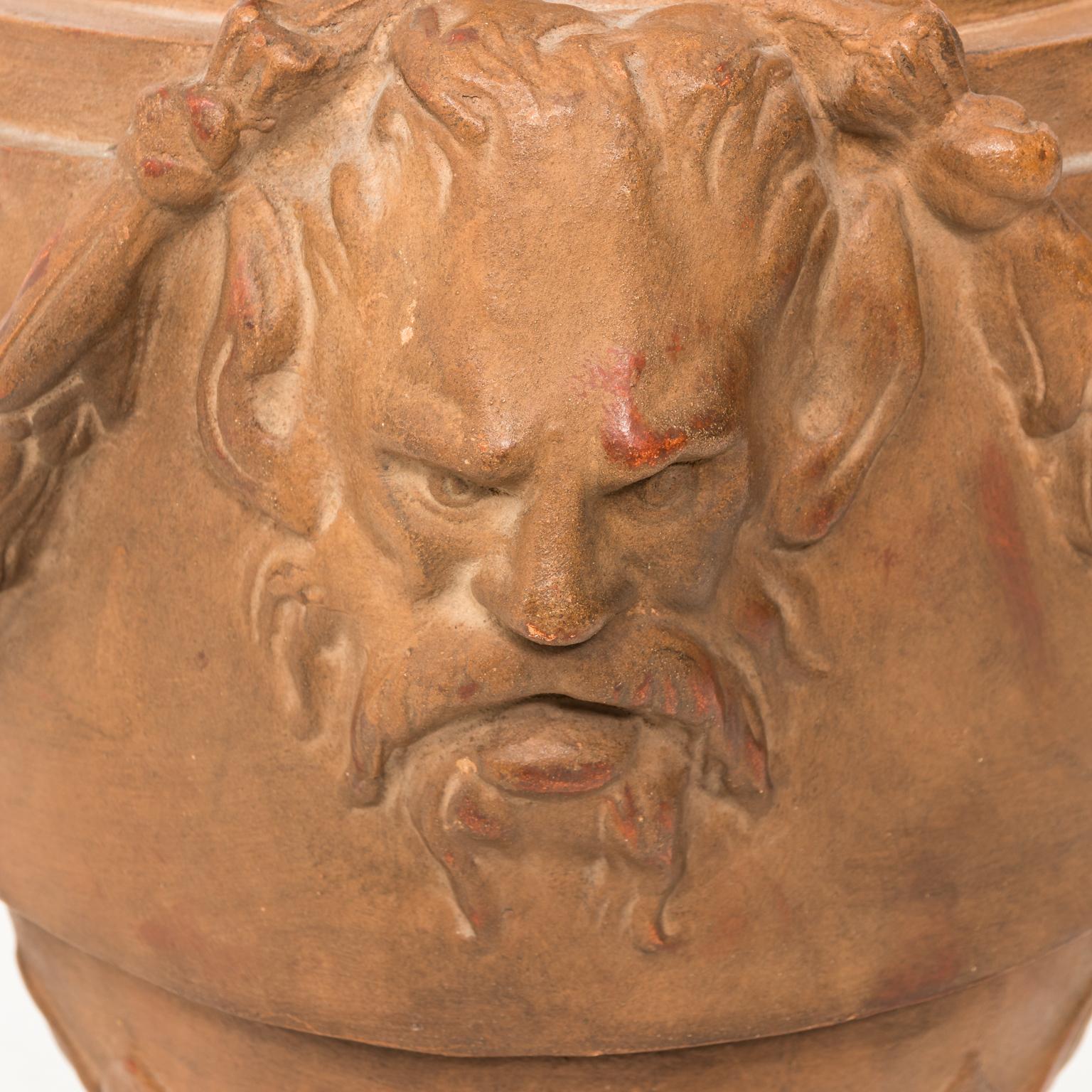 Neoclassical style terracotta urn with carved satyr heads and swags coming down the sides, resting on a square plinth. Hand-sculpted.
 