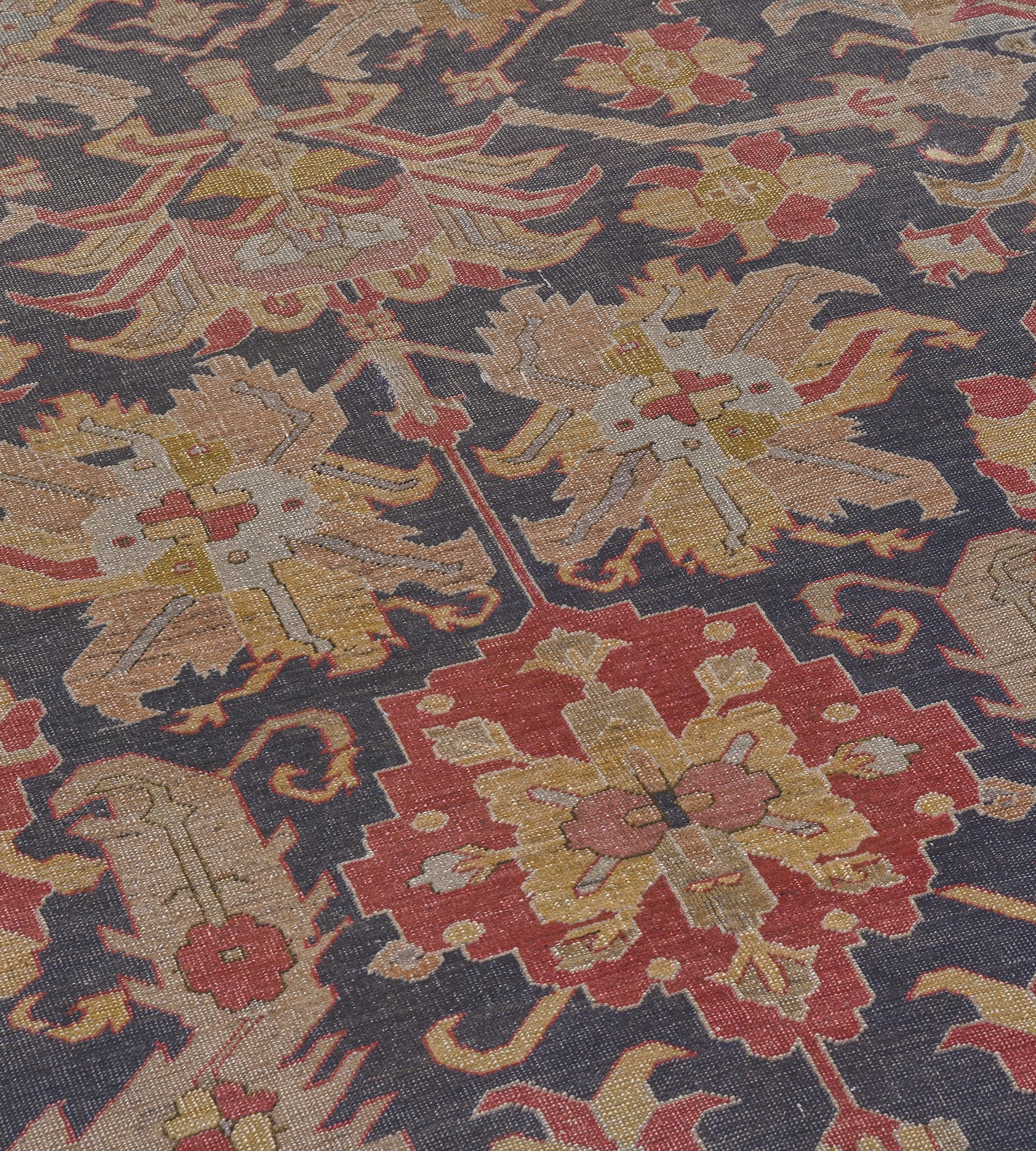 This traditional handwoven Turkish Hereke rug has a shaded indigo field with a central column of golden-yellow and brick-red linked palmettes and paired flowerheads surrounded by angular floral bouquets, flowerheads and serrated leaves, in a