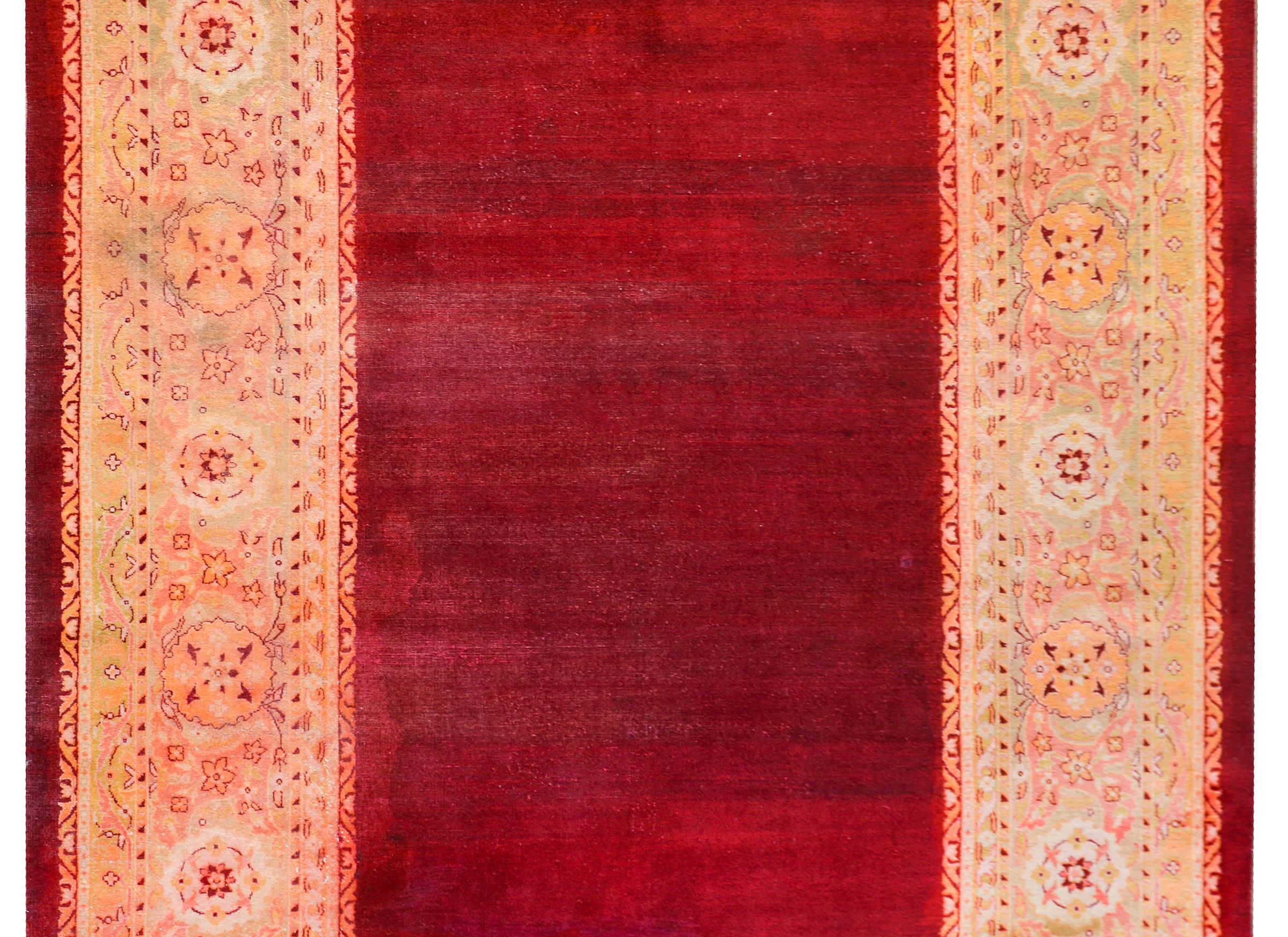 A beautiful early 20th century Indian Agra rug with an abrash crimson field surrounded by a wide floral pattered border woven in pale greens, golds, oranges, and pinks.