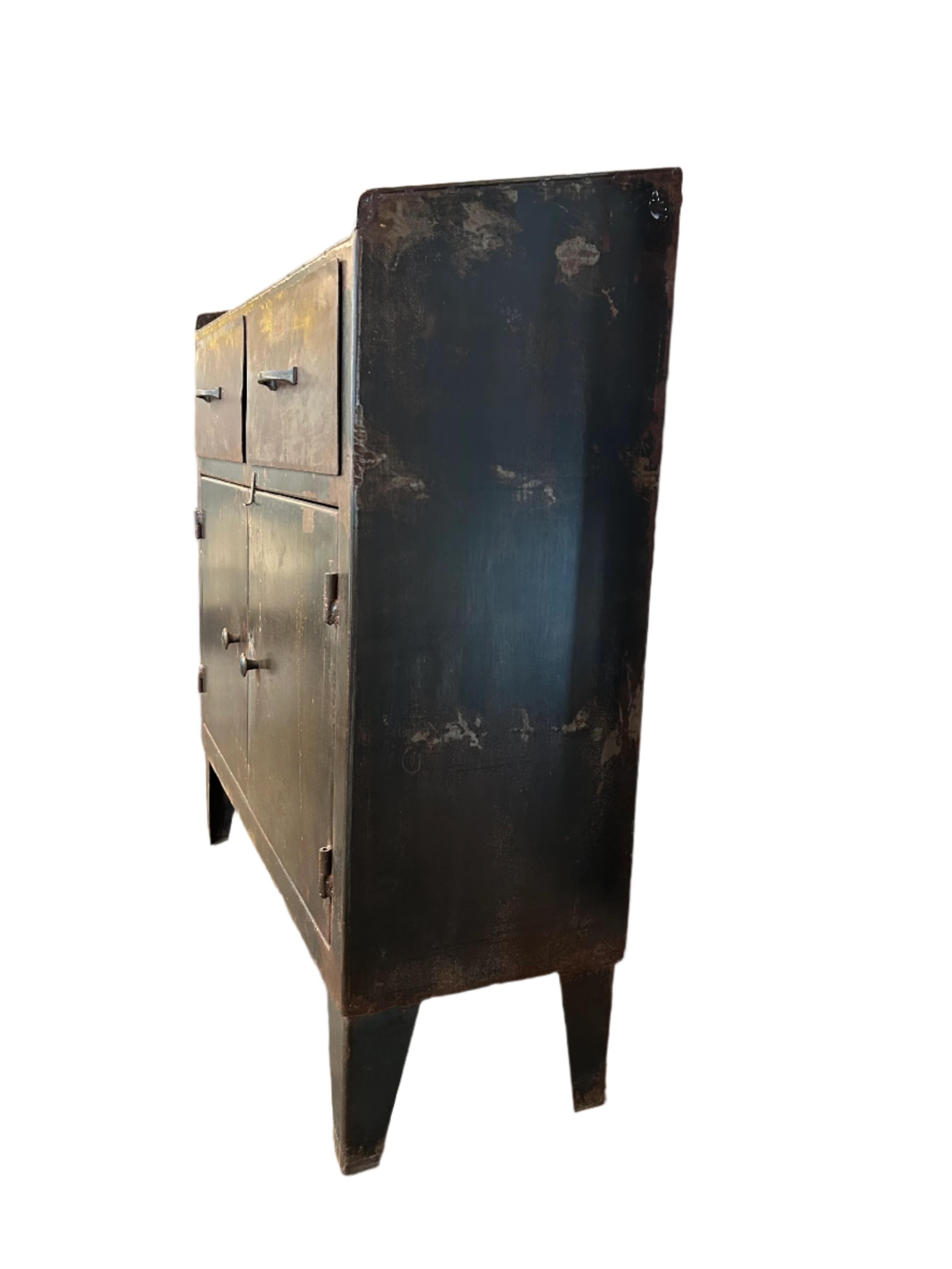 Esempio lampante di design utilitaristico, questa credenza industriale dei primi del Novecento mostra un carattere grezzo e una materialità onesta. Realizzato interamente in metallo, il pezzo presenta due profondi cassetti sopra un armadietto a due