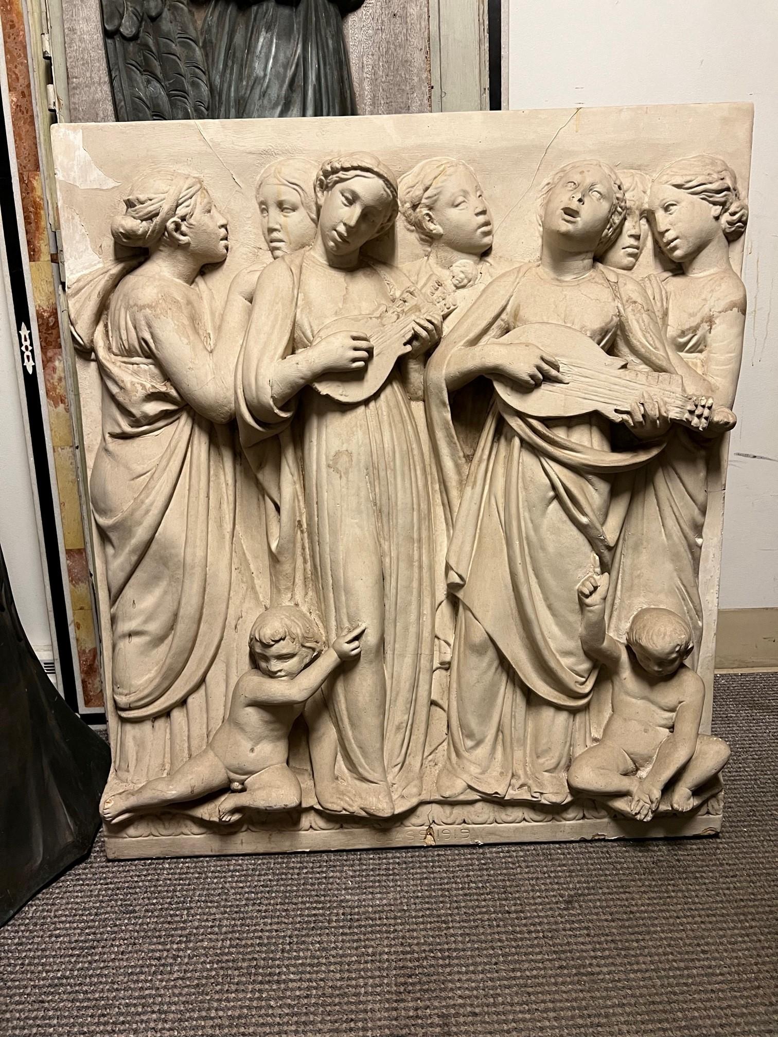 This is a large, high-relief wall hanging in early 20th century plaster. It depicts seven of the mortal daughters of Jupiter. They are singing, while two of them play stringed instruments, and they are standing above two young boys who are