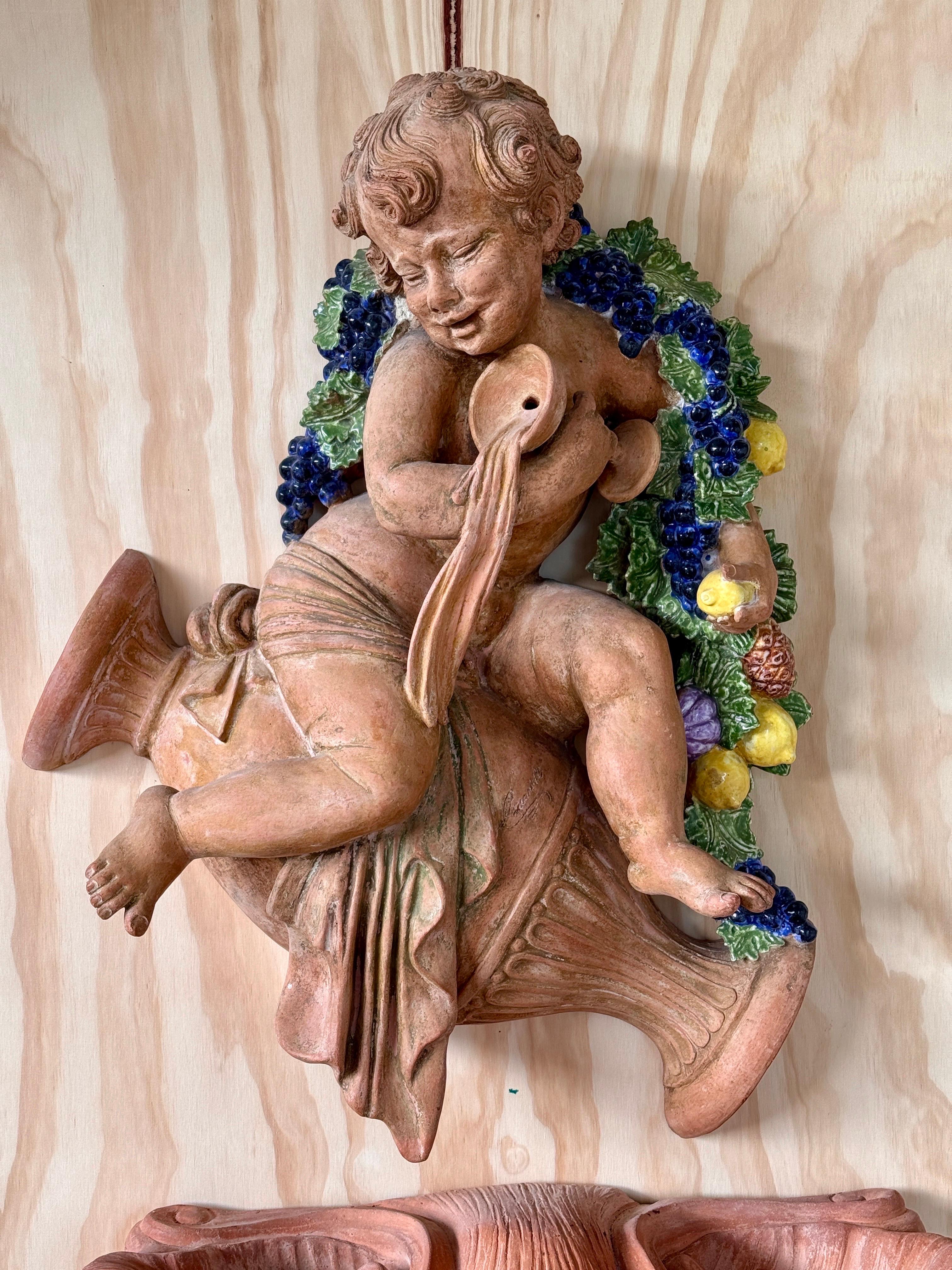 A striking early 20th-century Italian terracotta wall fountain featuring Bacchus, the Roman god of wine and revelry.
This two-piece set includes a sculptural relief of a cherubic Bacchus draped in flowing fabric, adorned with a garland of grapes and