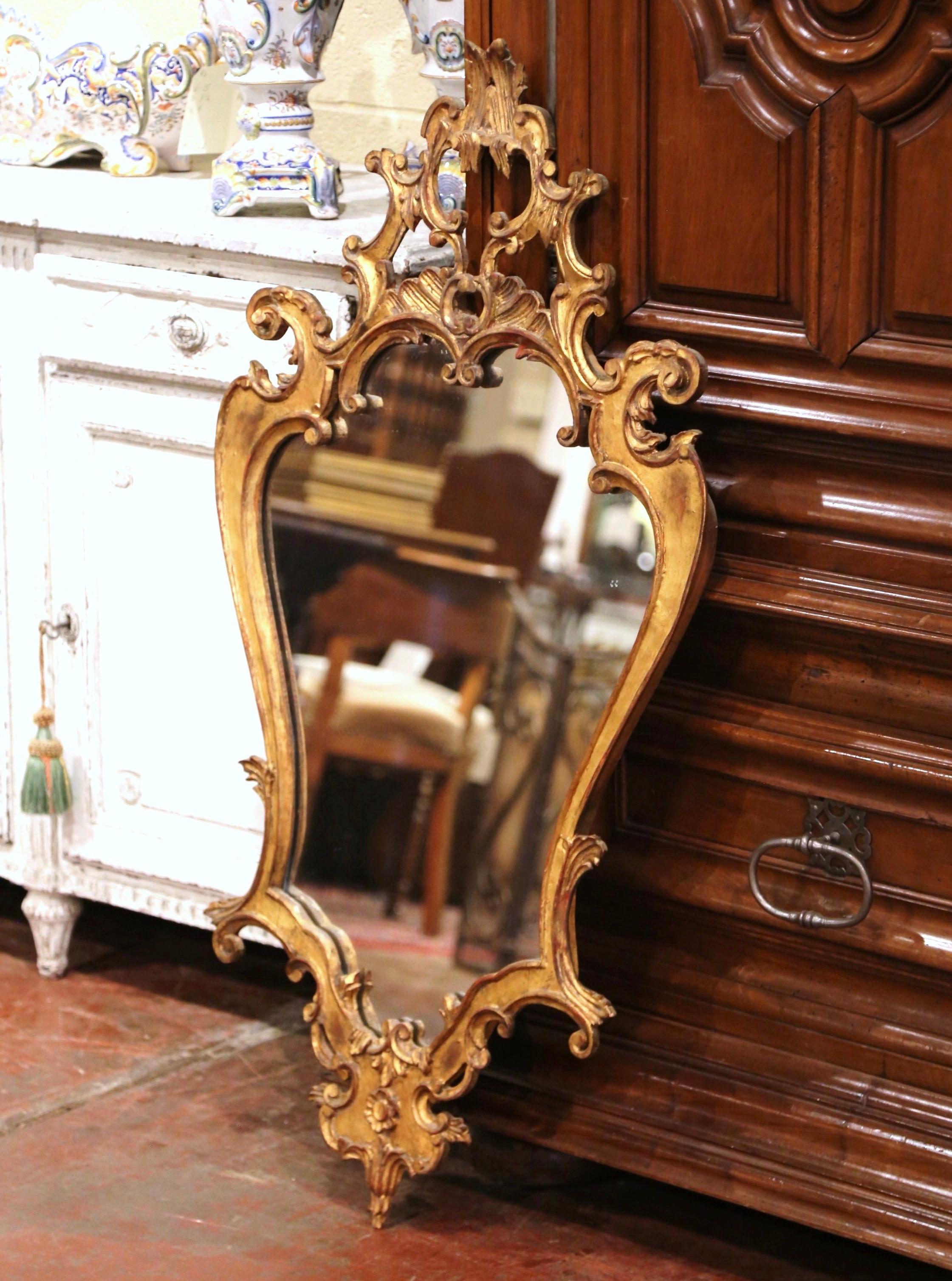 Crafted in Italy, circa 1920, the gilt mirror in the Louis XV style, features intricate carvings, including scroll and floral decor, and embellished with a carved shell cartouche at the top and bottom. The Rococo mirror is in excellent condition and