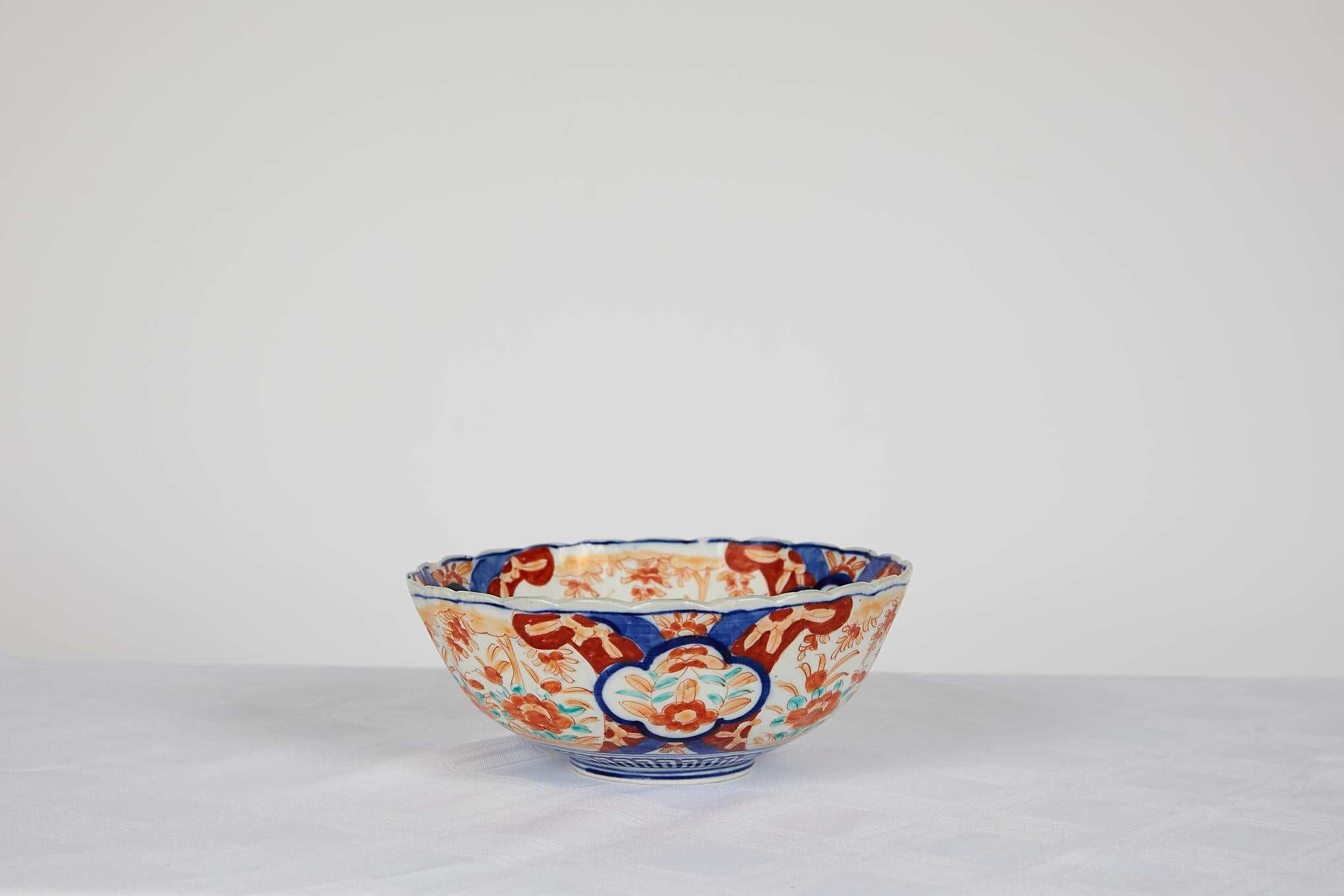 Early 20th century Japanese Imari scalloped bowl with a center medallion surrounded by alternating floral decorations hand painted in hues of cobalt blue, burnt orange, and emerald green.
