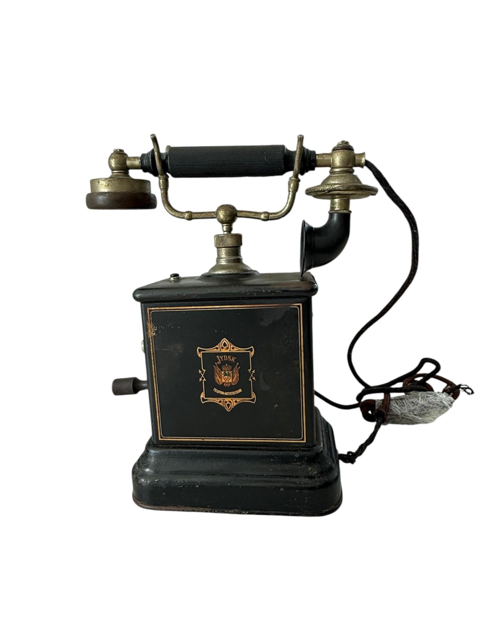 Il s'agit d'un téléphone de bureau Jydsk du Danemark datant du début du 20e siècle. Elle représente une boîte métallique carrée de couleur noire, avec un téléphone à manivelle de bureau. Sur la face avant du téléphone figure le Label de l'entreprise