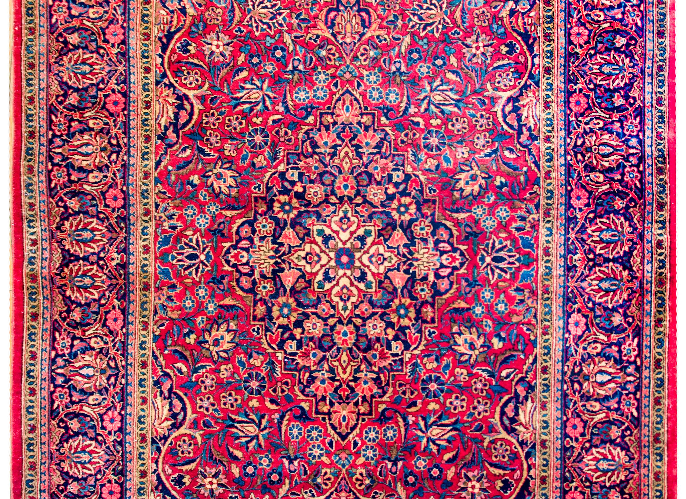 A gorgeous early 20th century Persian Kashan rug with an all-over pattern of myriad flowers and scrolling vines woven in light and dark indigo, cream, pink, and crimson on a rich crimson background. The border is extraordinary with a wide central