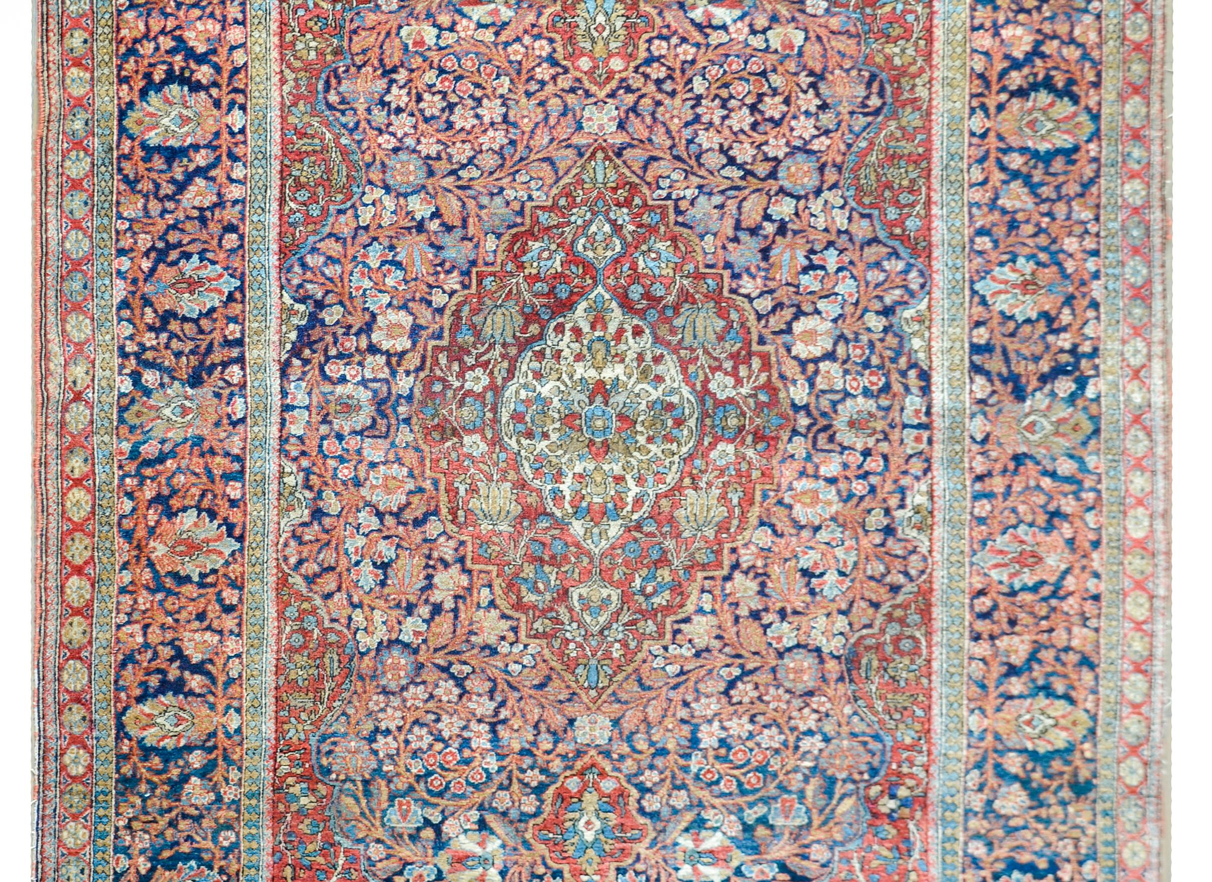 A fantastic early 20th century Persian Kashan rug with a mesmerizing pattern with a large central medallion filled with myriad flowers, and living among a field of more flowers and scrolling vines, and all woven in crimson, pink, gold, and cream