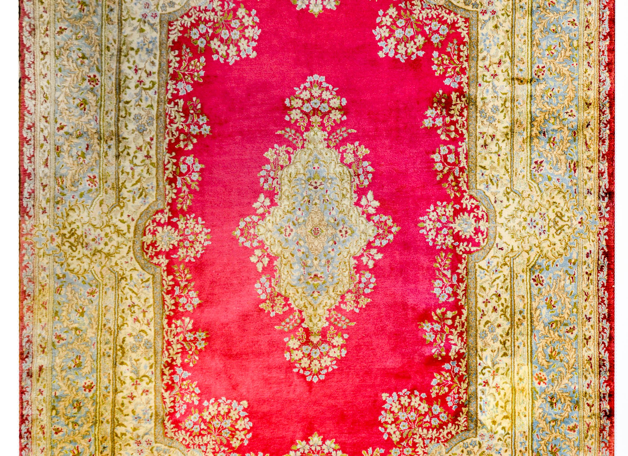 A circa 1950 Persian Kirman rug with a large central floral medallion with myriad flowers and vines woven in taupe, green, pale blue, on a bold cranberry colored background. The border is wide and of a matching pattern and colors as the medallion.