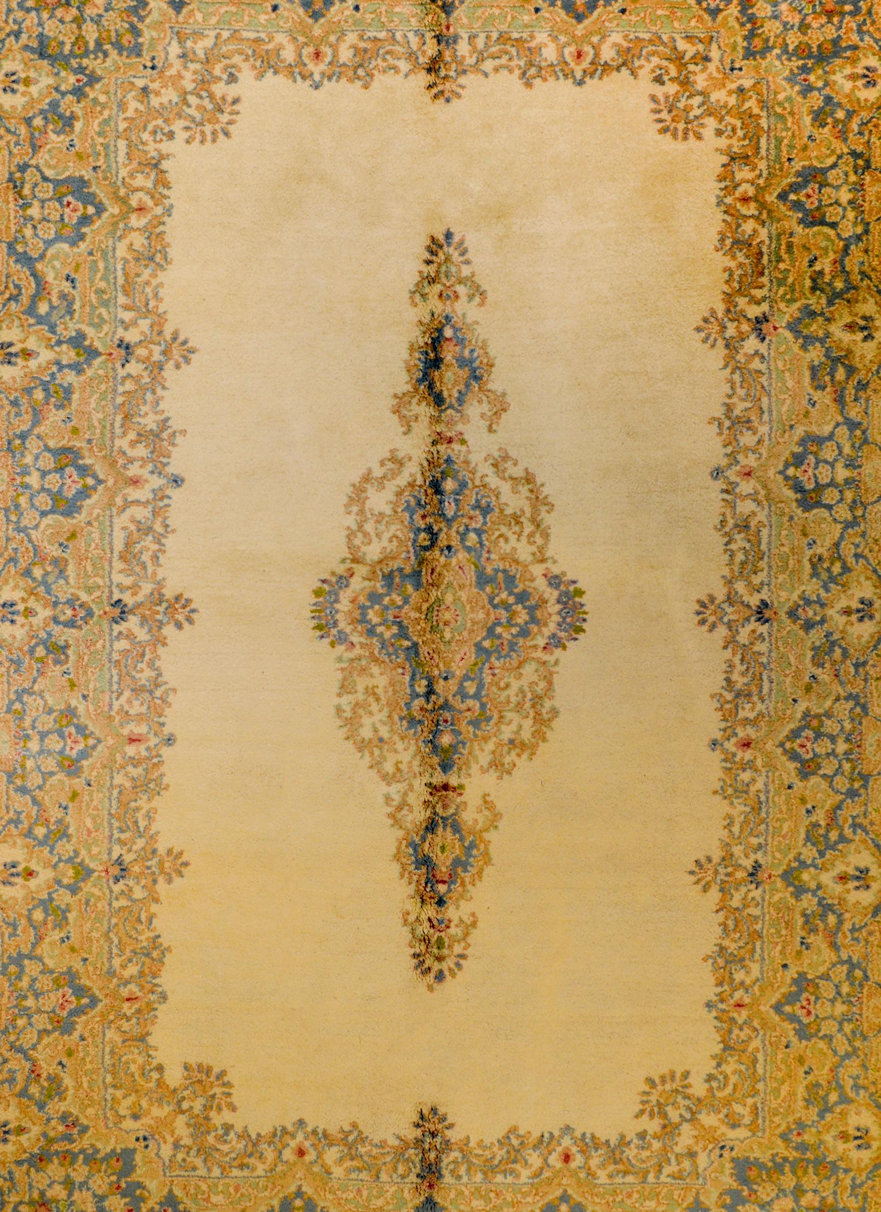 An unusual early 20th century Persian Kirman rug with a long narrow floral medallion woven in indigo, green, gold, and red vegetable dyed wool on a champagne colored ground. The border is wide with a similarly styled pattern as the medallion, but