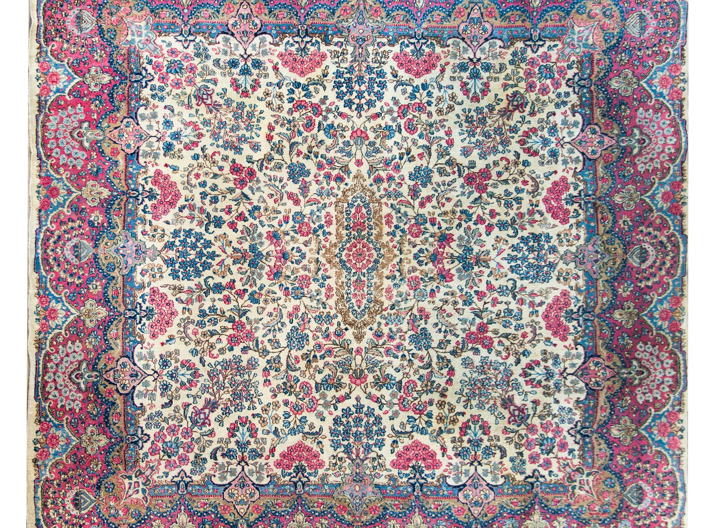 A stunning early 20th century Persian Kirman rug with a beautiful mirrored floral pattern containing myriad floral clusters woven in pinks, indigos, and greens, and set against a cream colors background, and all framed by a wide floral pattered