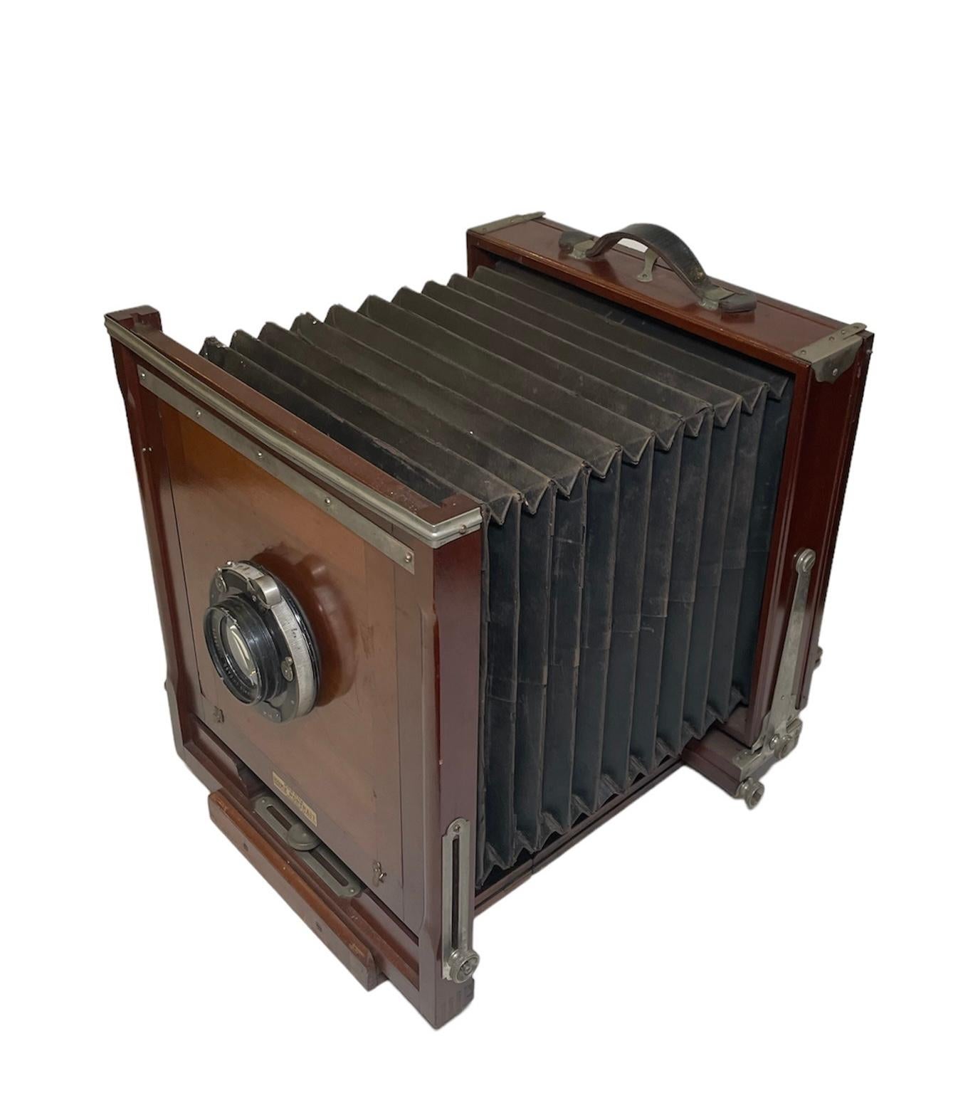This is a Korona Home Portrait Camera. It depicts a square wood camera body and base with square glass in the back and metal hardware. In the middle, there is an accordion like black leather structure. A black leather handle is in the top center. A