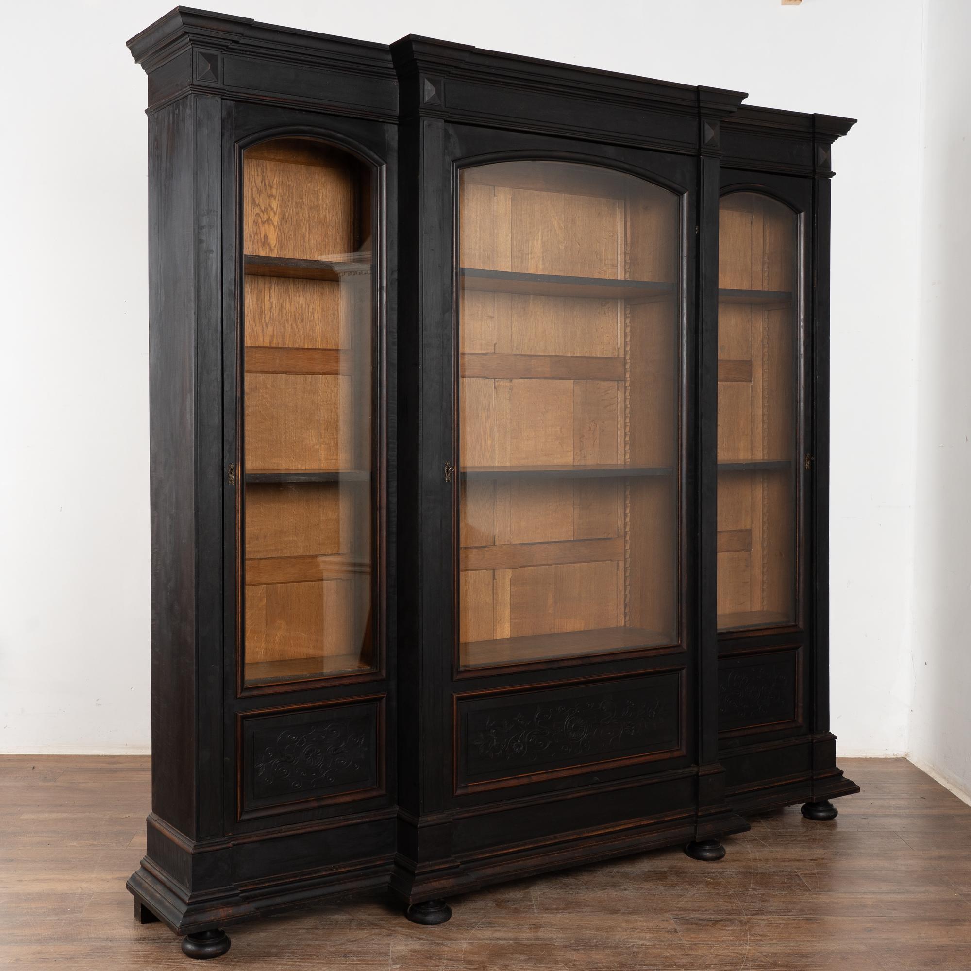 This large oak bookcase or display cabinet has three long glass doors and stands over 7.5' tall and wide. Carved details embellish the lower panels.
Later professionally painted in black, creating a fresh and dramatic appeal to this striking