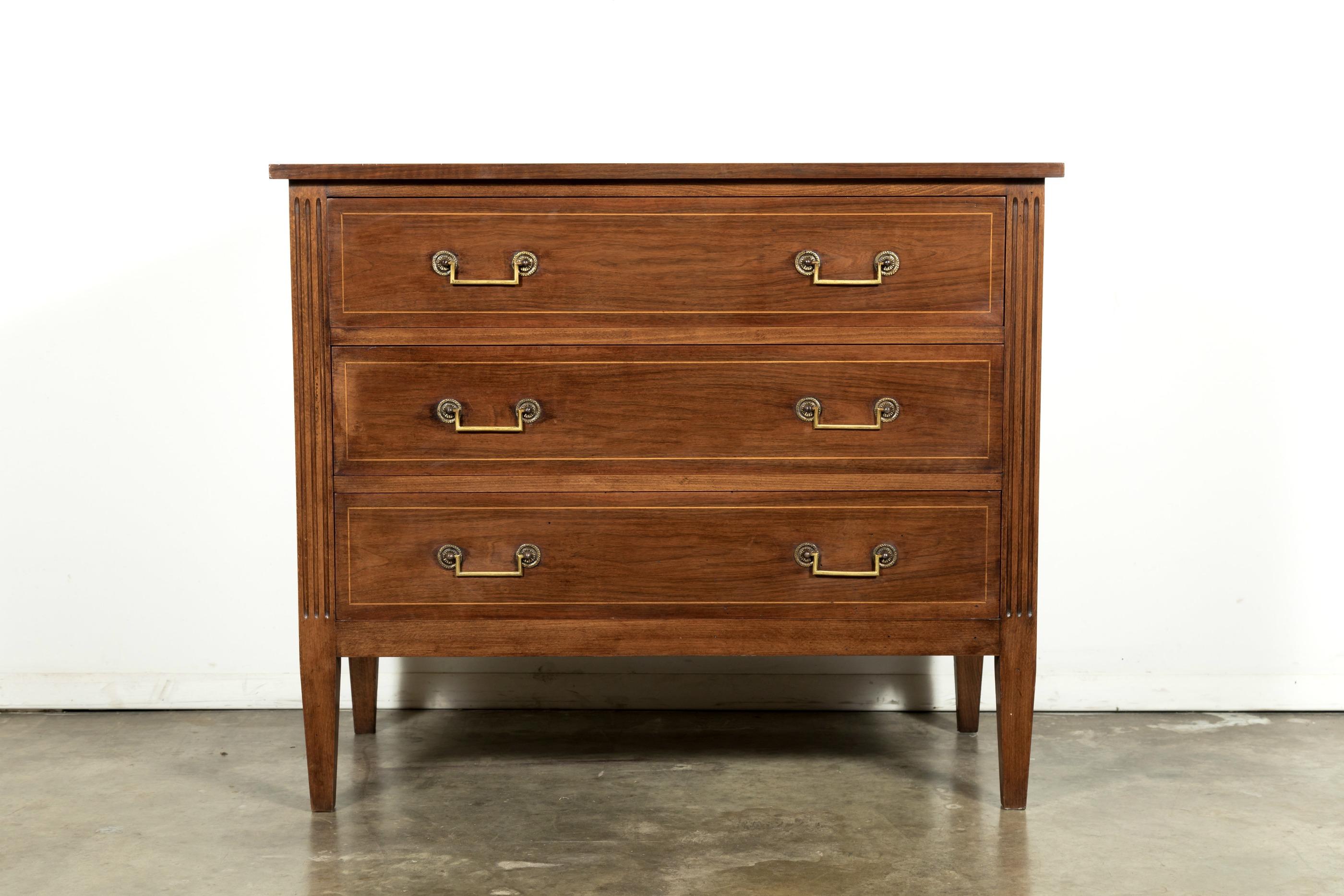 A handsome early 20th century Louis XVI style Parisian commode handcrafted of walnut and fruitwood parquetry, having three drawers adorned with original brass drop bail handles, supported on fluted stiles, sitting on short tapered legs. This simple,