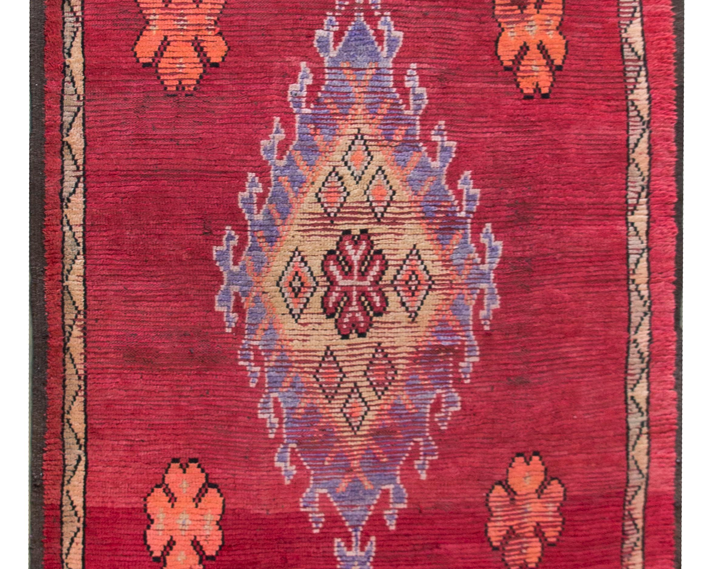 A stunning early 20th century Moroccan Berber runner with a large central diamond medallion amidst a rich crimson field of more stylized flowers woven in orange, gold, violet, and pink, and surrounded by a thin geometric patterned stripe.