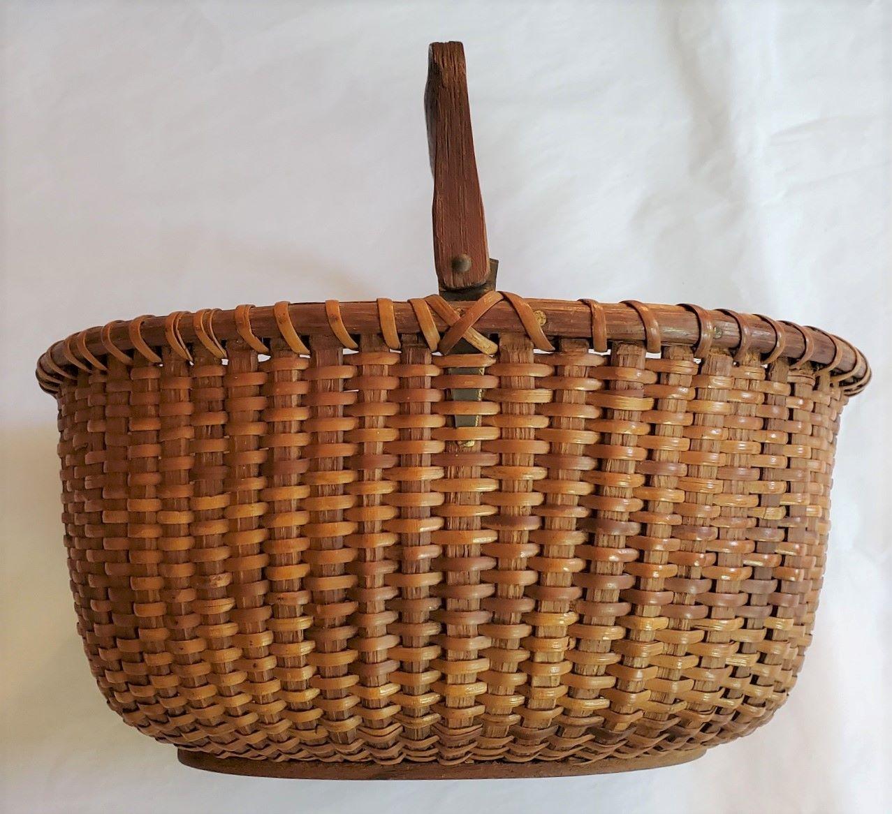 Early 20th Century Nantucket Oval Basket attributed to A.D. Williams (Nantucket: 1867 - 1940), circa 1920, an oval open swing handled Nantucket basket with cane weave on oak staves, slender carved handle attached to short tin ears entrapped under