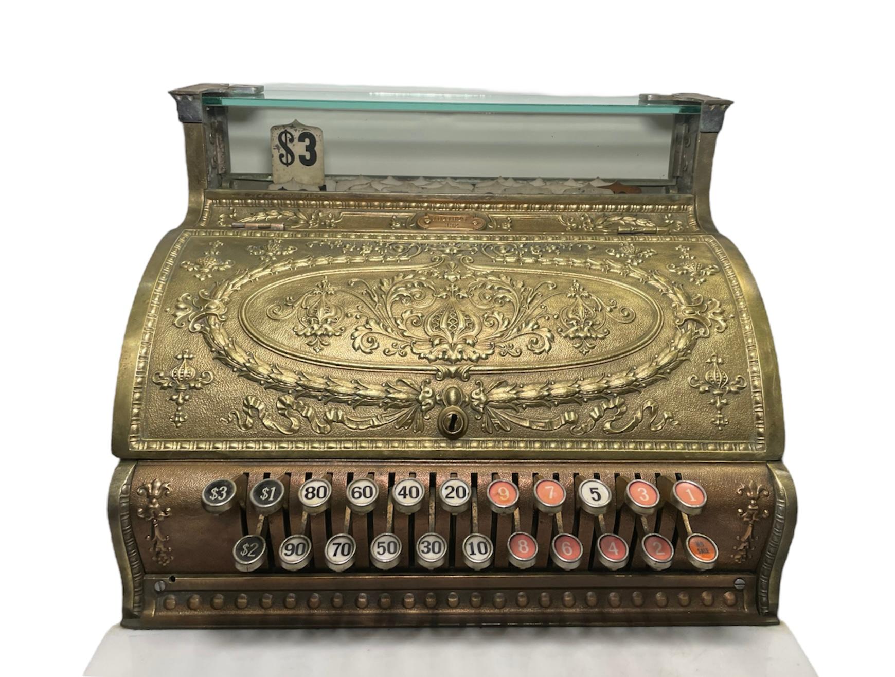 This is a gilt brass heavy manual National Cash Register model 332. It is embellished with the embossing of small and large onion shaped flowers, wreaths of bay leaves, scrolls of acanthus leaves and garlands of flowers with bows/ribbons. At the