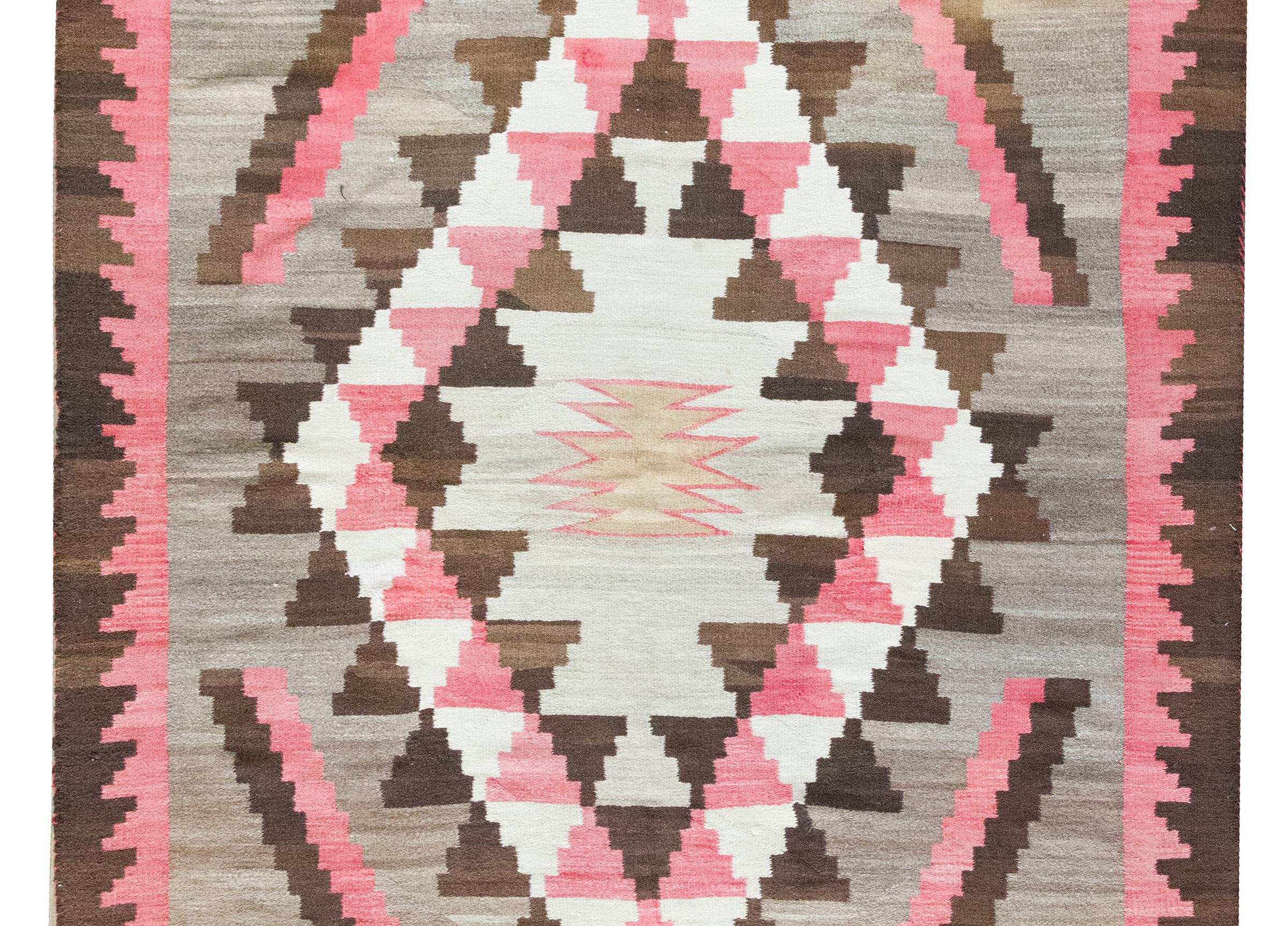 Woven in the early 20th century, this striking Navajo rug features a bold central zigzag medallion rendered in a soft crimson stripe, enclosing a calm gray field. The medallion sits within a larger diamond form composed of repeated triangular motifs