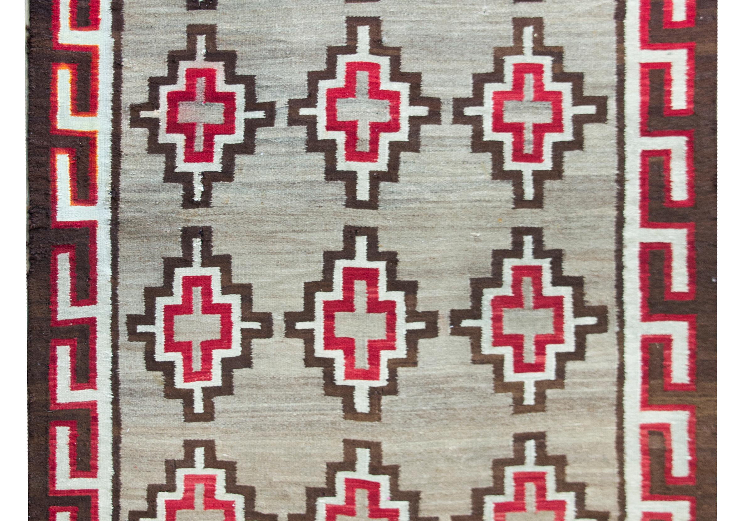 A striking early 20th century Navajo rug with an all-over stepped diamond pattern with crimson, white, and black stripes, set against an abrash natural gray wool ground, and surrounded by a meandering motif border.