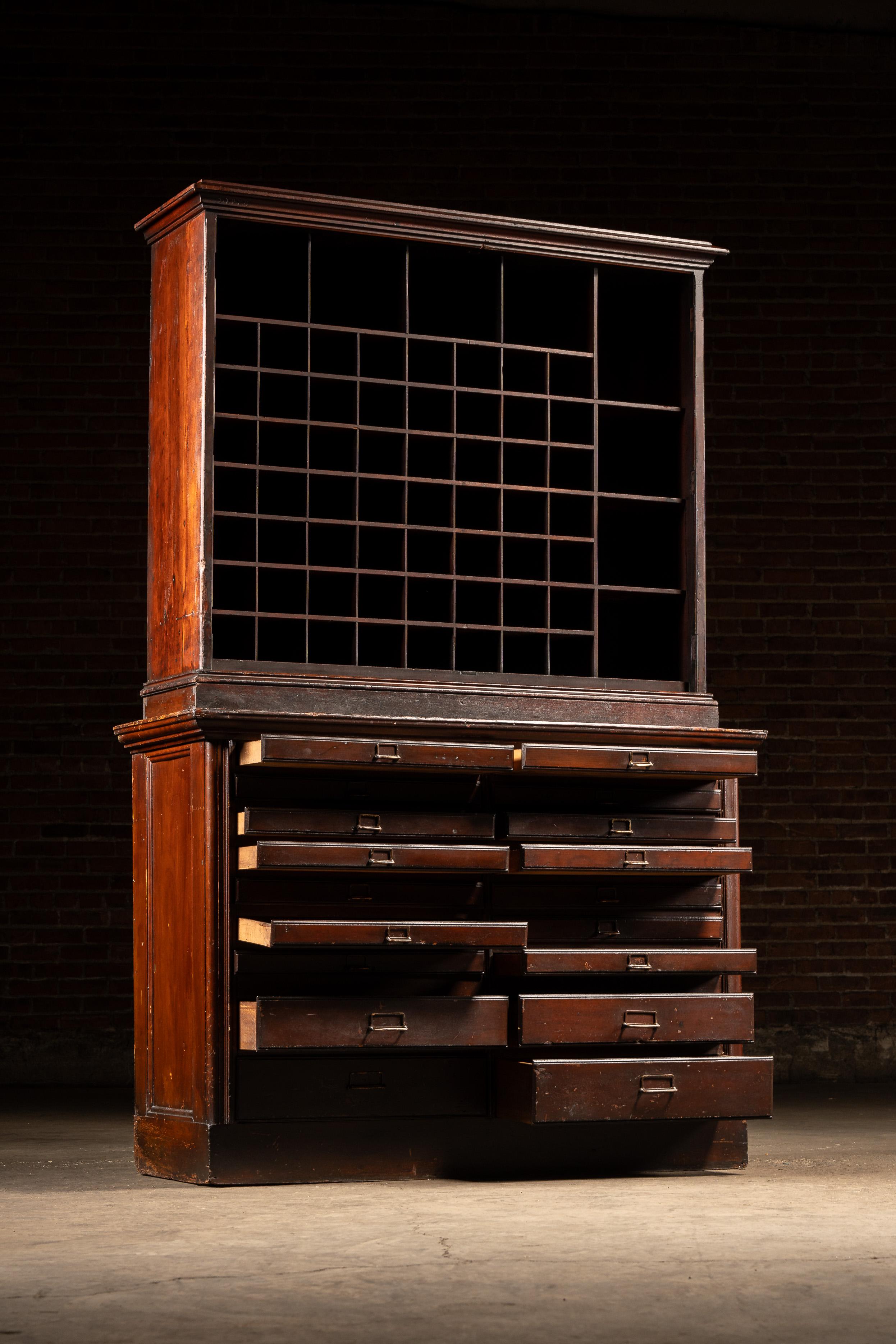 Large two-piece oak cabinet featuring a double-bank of flat file drawers below and a gridded cubby organizer above. The upper section includes dozens of pigeonholes for sorting mail or documents, while the base houses nine graduated drawers per