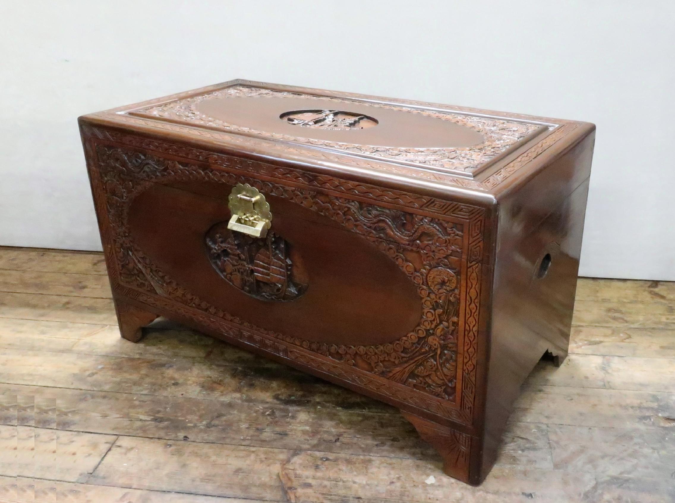 A decorative early 20th century oriental freestanding teak chest with carvings of dragons, boats and pagodas to the front and top stood on bracket feet. The chest retains its original engraved brass back plate and latch and comes with a lock and