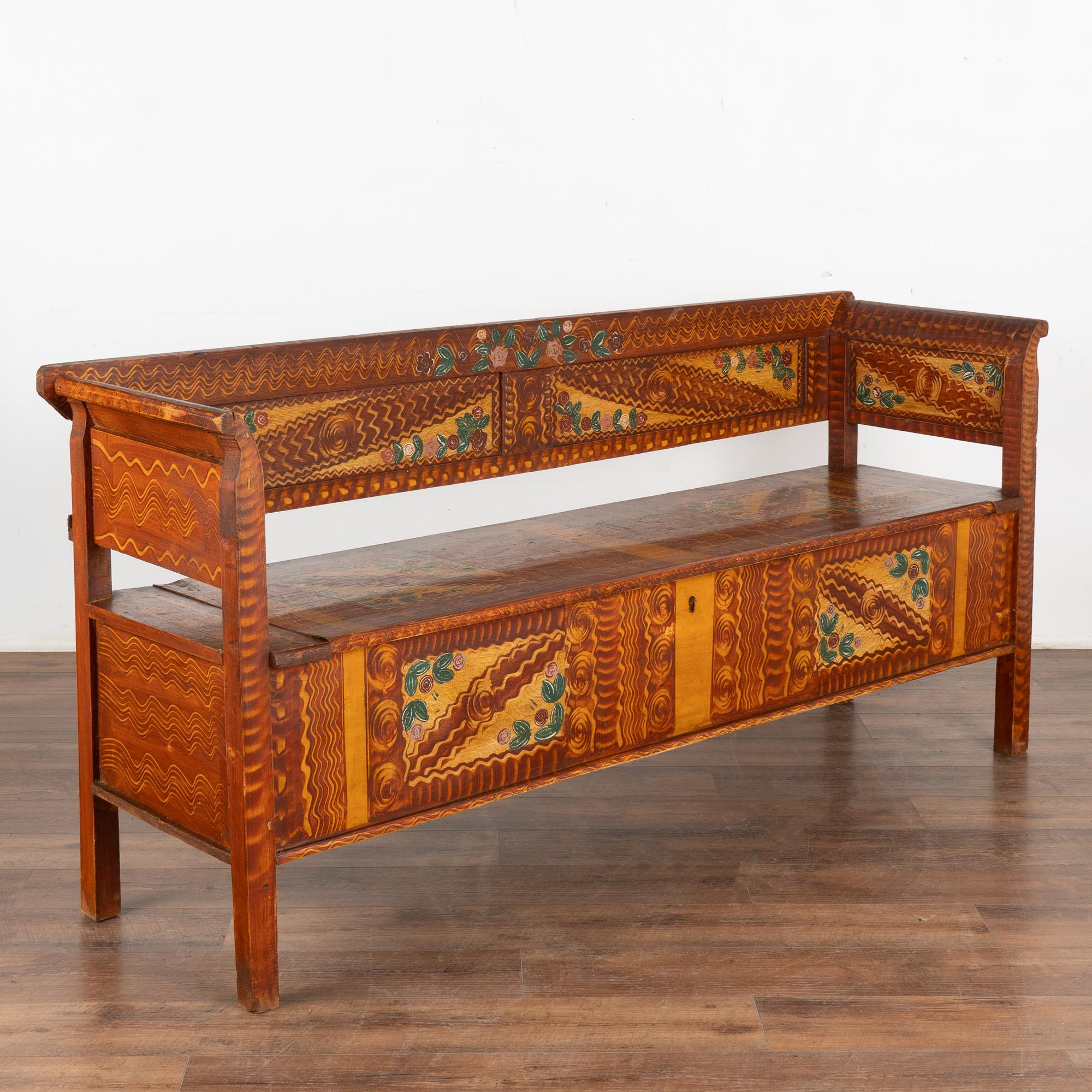Ce banc original en pin, peint à la main, présente un fond stylisé en faux bois brun et un motif floral coloré.
La banquette traditionnelle à charnières révèle les rangements intérieurs. 
Restauré, nettoyé et ciré, ce banc de 6,5 pieds de long est