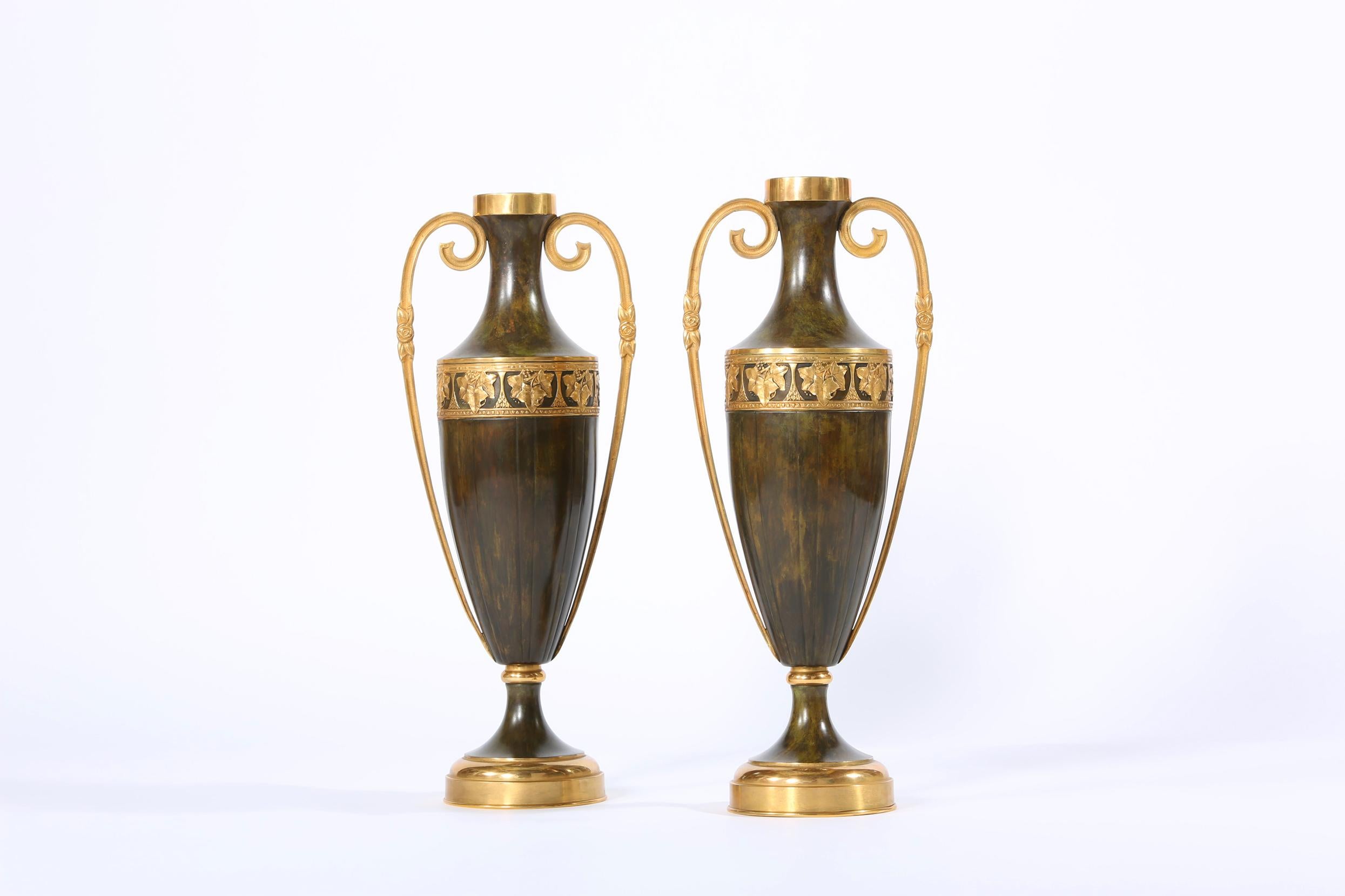 Early 20th century pair of tall decorative patinated bronze vases / pieces with exterior design details and gilded side handles. Each vase is in great vintage condition. Minor wear consistent with age / use. Each piece stand about 22.5 inches tall x