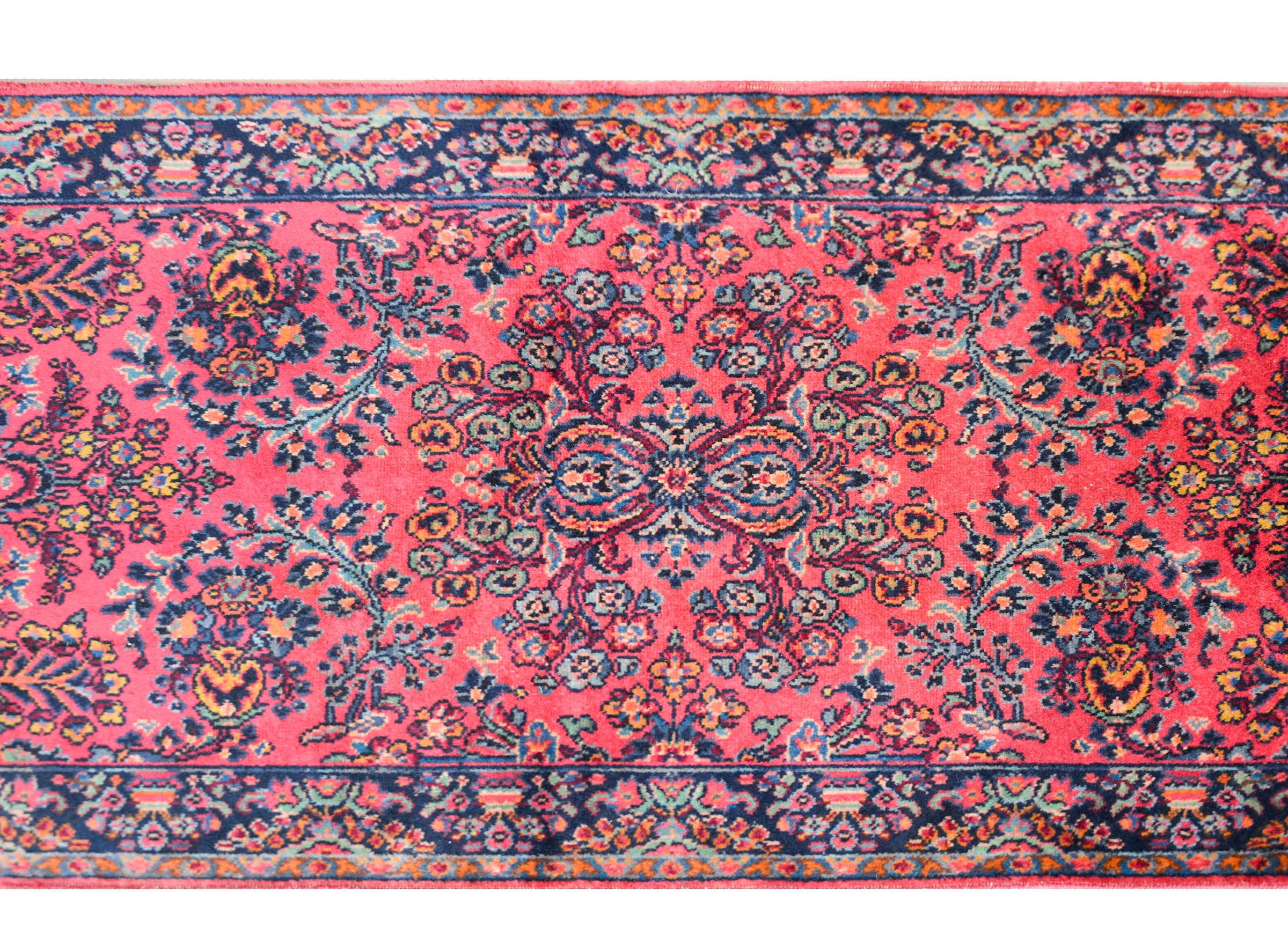 An early 20th century Persian Karastan Sarouk patterned rug with an all-over floral and leaf pattern woven in light and dark indigo, gold, teal, and all set against a pink background. The border is sweet with a floral and tree-of-life pattern woven