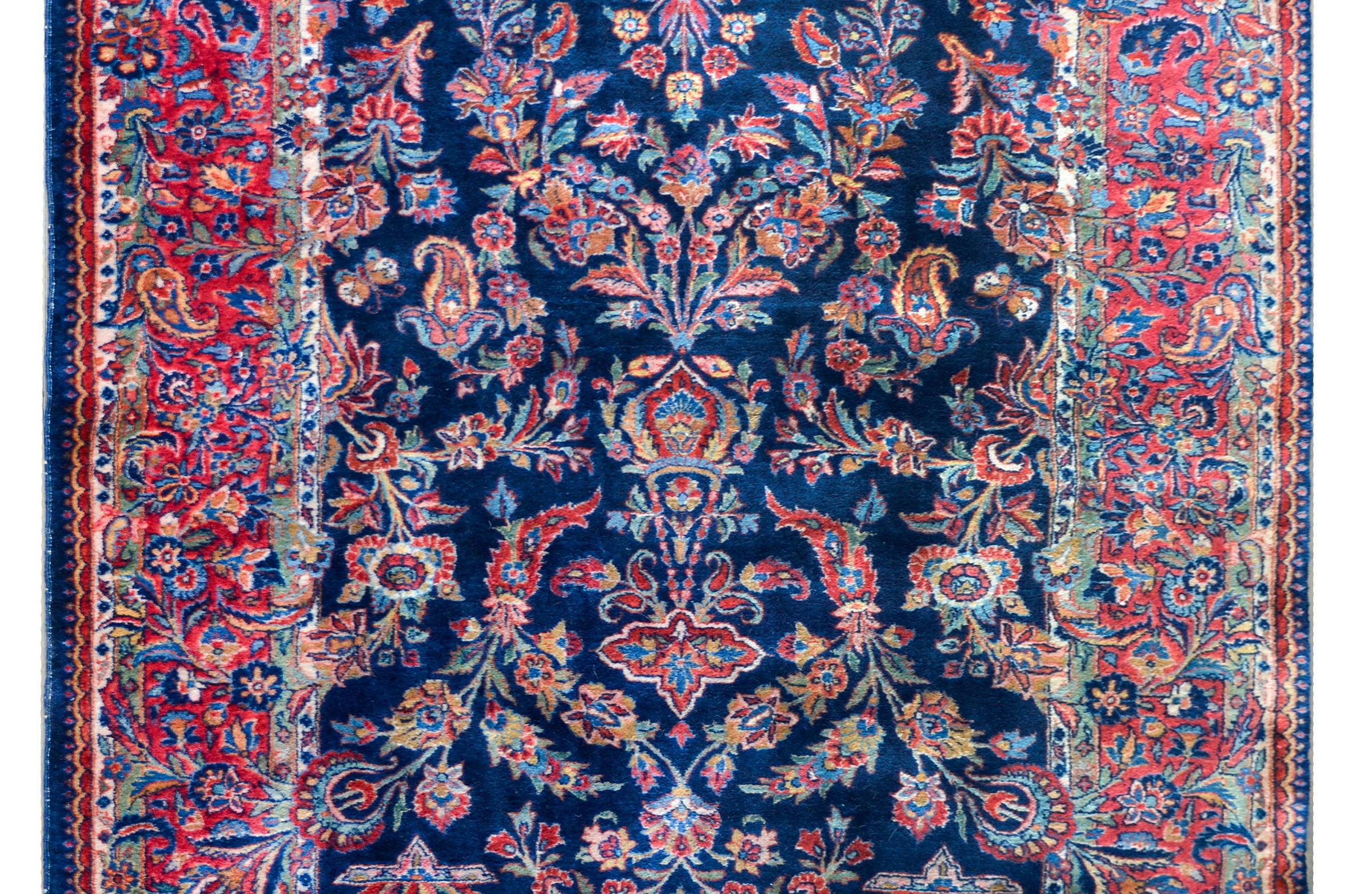 An early 20th century Persian Kashan rug with a mirrored pattern containing myriad flowers woven in reds, blues, golds, and greens, and set against a dark indigo background. The border is fantastic composed with more flowers woven in similar colors