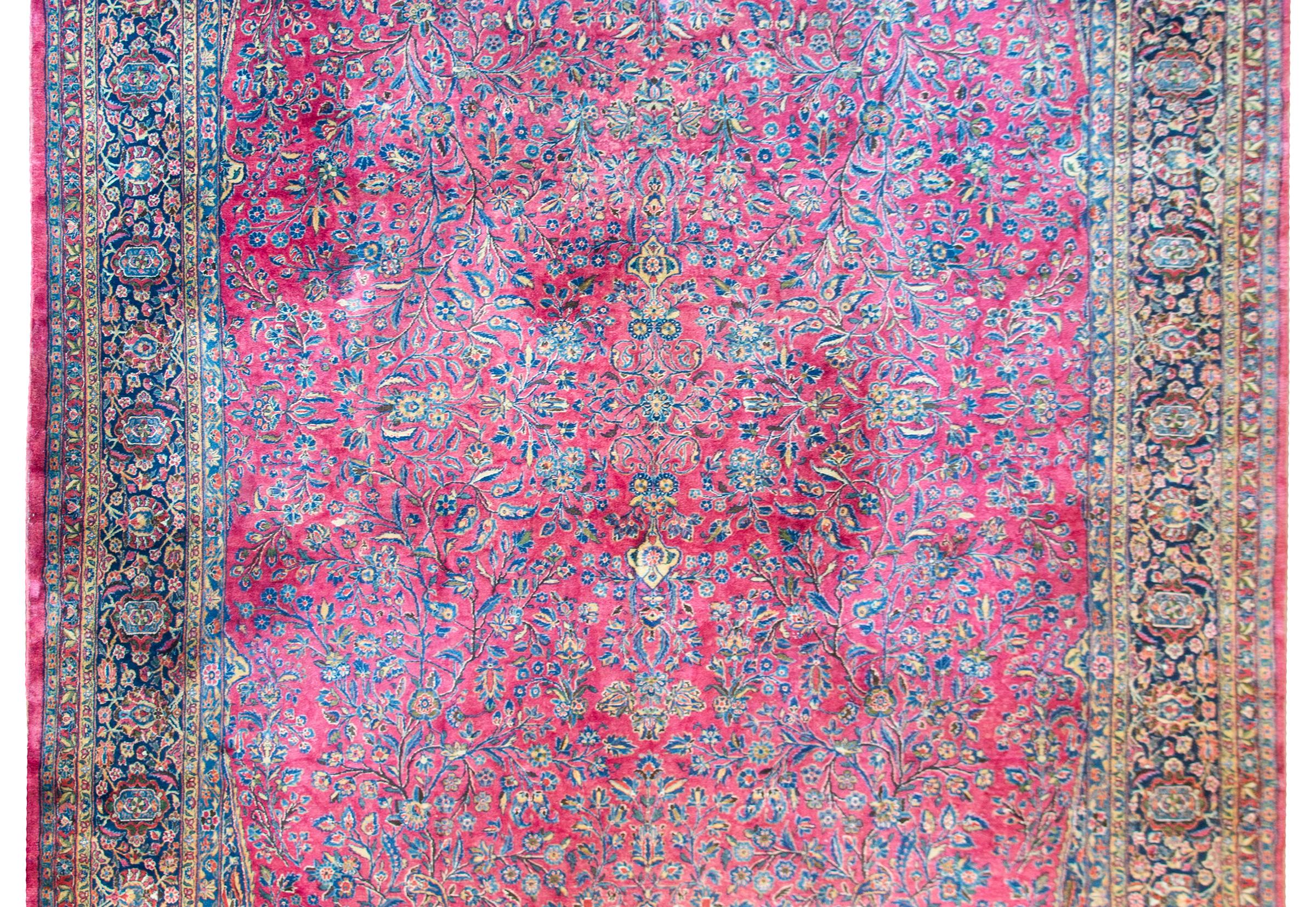 A wonderful early 20th century Persian Kashan rug with an all-over floral and scrolling vine pattern woven in indigos, creams, and pinks, and set against a cranberry colored field, and all surrounded by a complex wide floral and scrolling vine