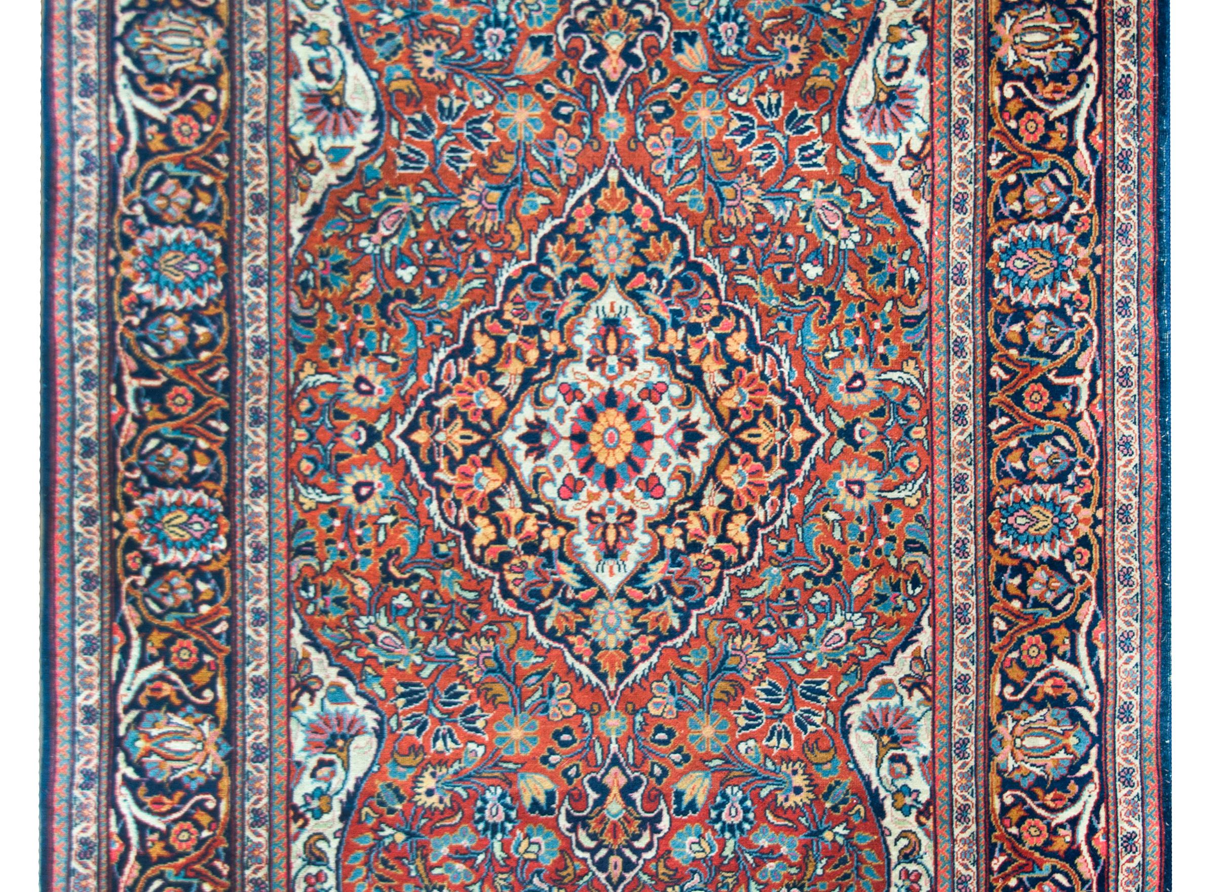 A playful early 20th century Persian Kashan rug with a wonderful central floral medallion woven in crimson, indigo, coral, and orange, all set against a floral and scrolling vine field against a crimson background. The border is stunning, with a
