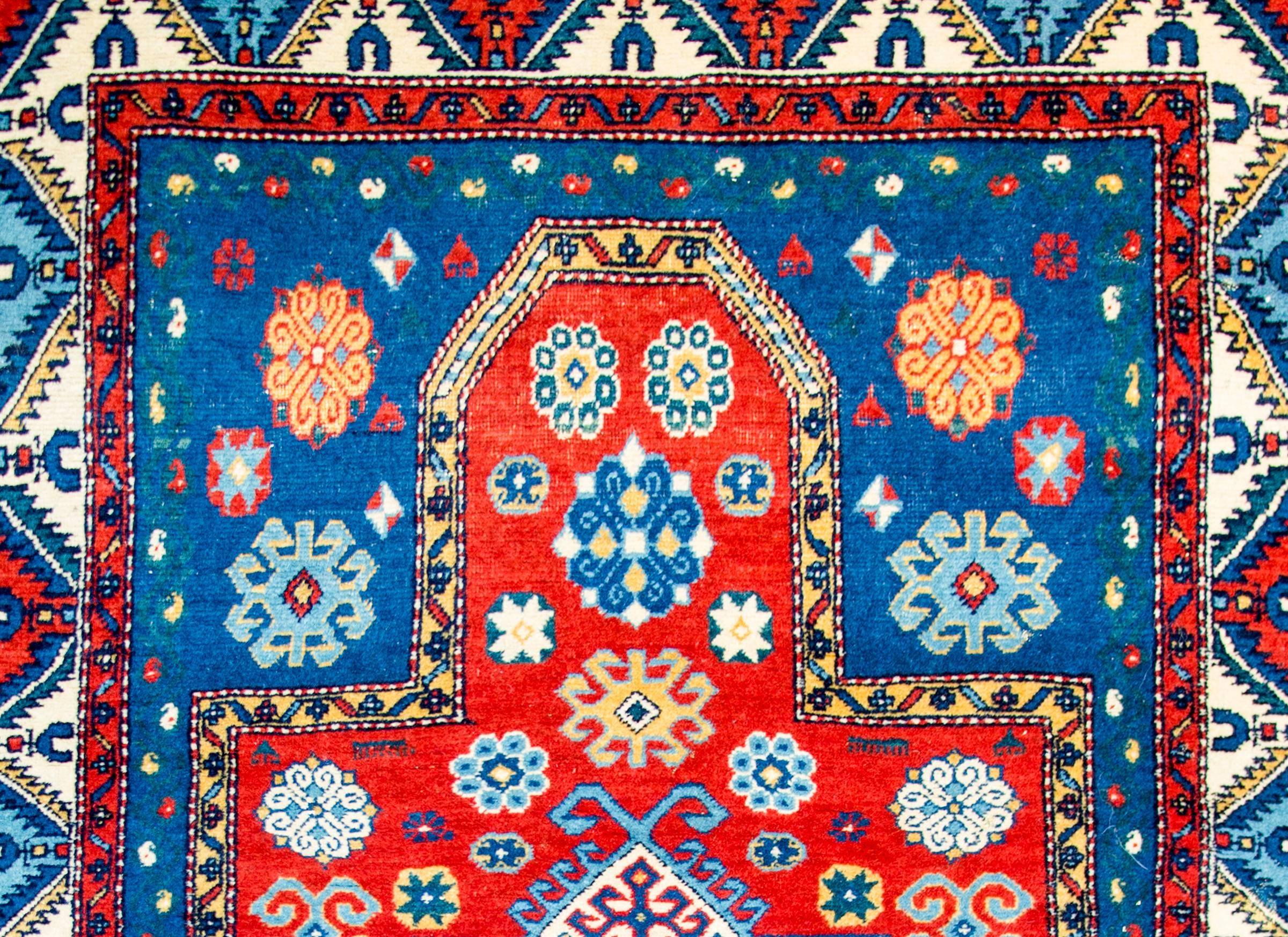 An early 20th century Kazak prayer rug with an all-over stylized floral pattern woven with light and dark indigo, and gold colored wool on a brilliant crimson colored background. The border is complex with multiple stylized petite floral patterned
