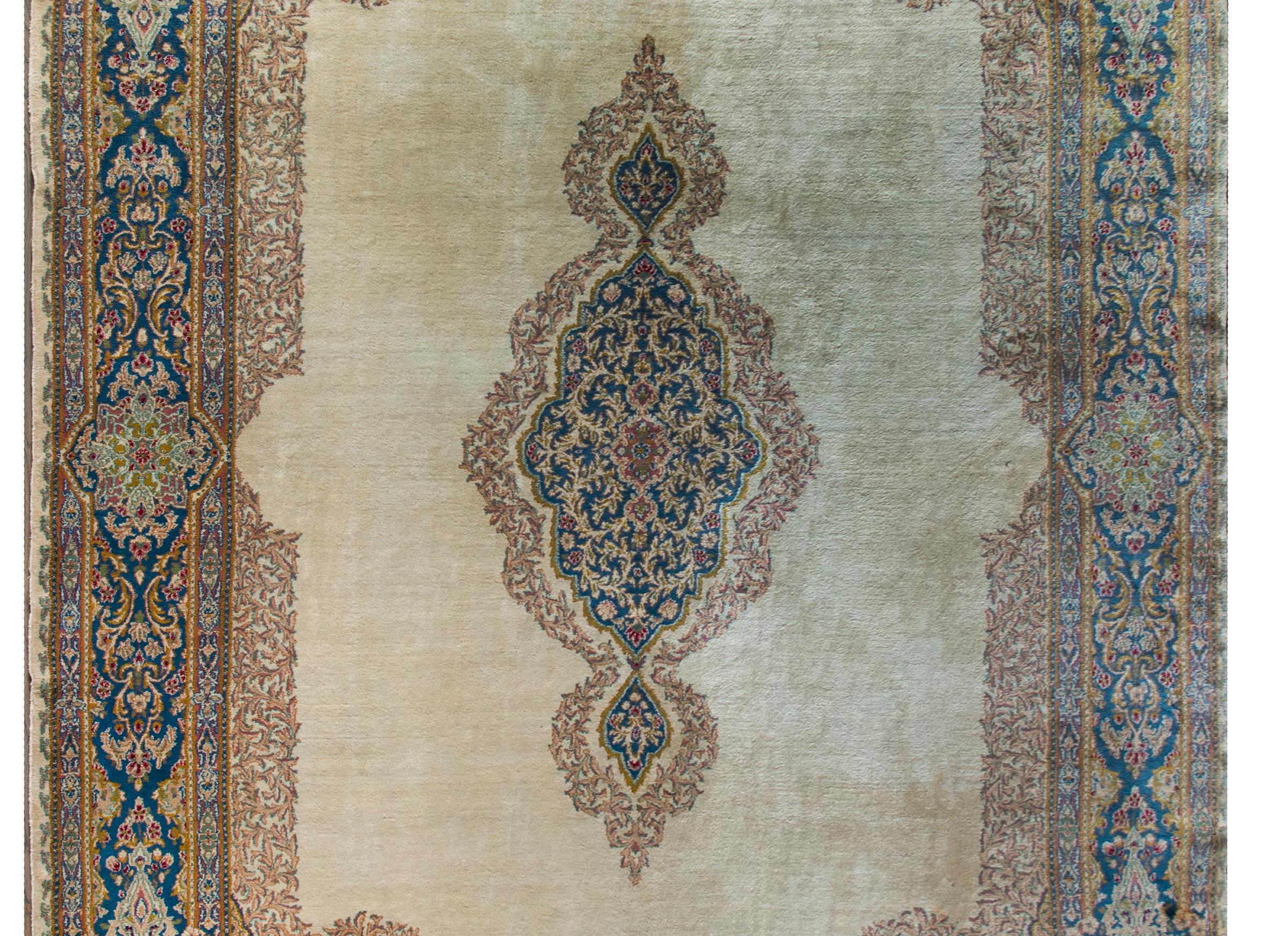 A beautiful early 20th century Persian Kirman rug with a wonderful central floral medallion woven in pink, orange, indigo, and cranberry and set against a stunning champagne colored field, and all surrounded by an extraordinary border containing