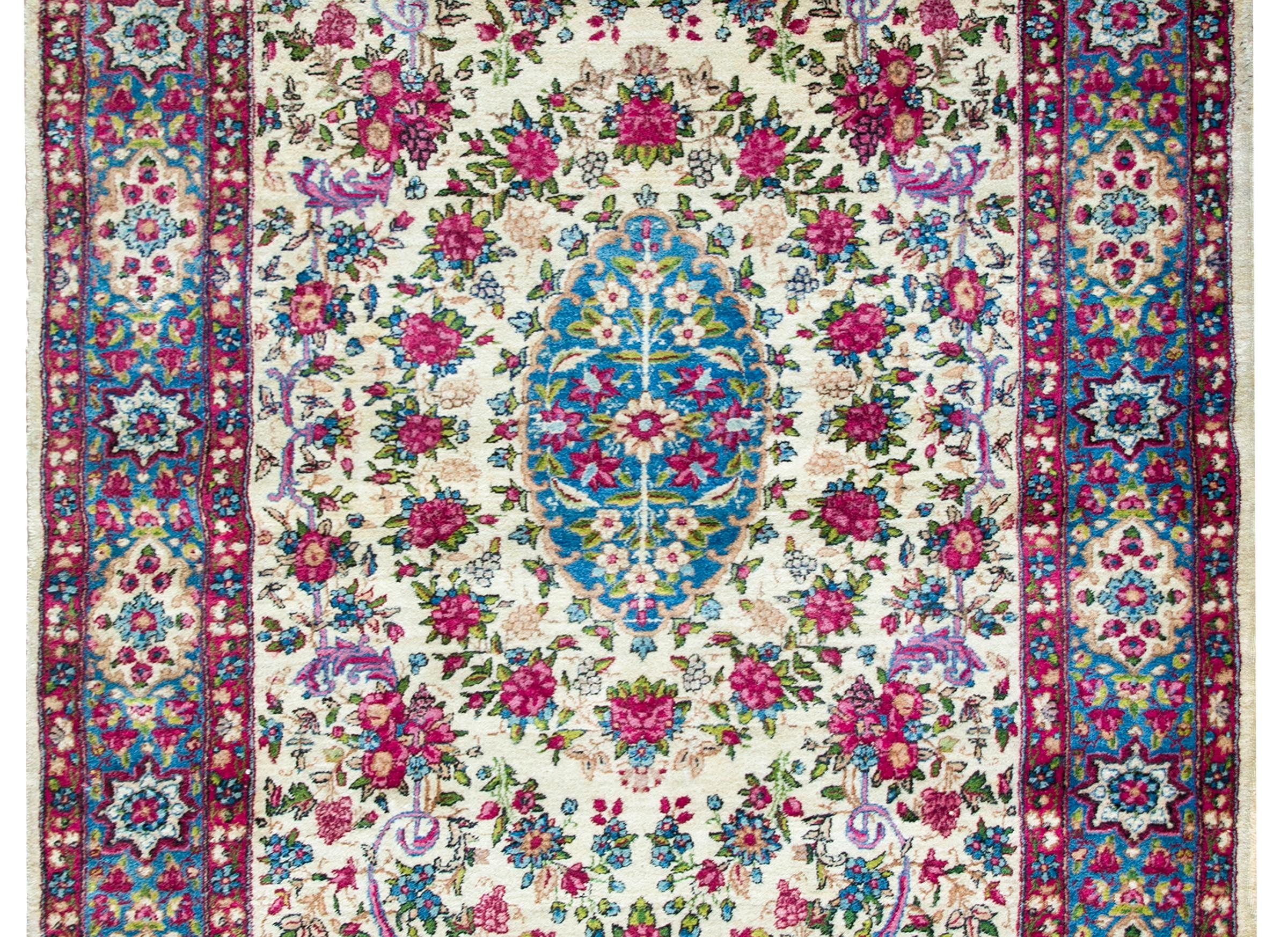 A sweet early 20th century Persian Kirman rug with a stunning pattern with myriad flowers woven in pinks, fuchsias, indigos and green, all set against a cream colors background, and surrounded by a complex stylized floral patterned border.