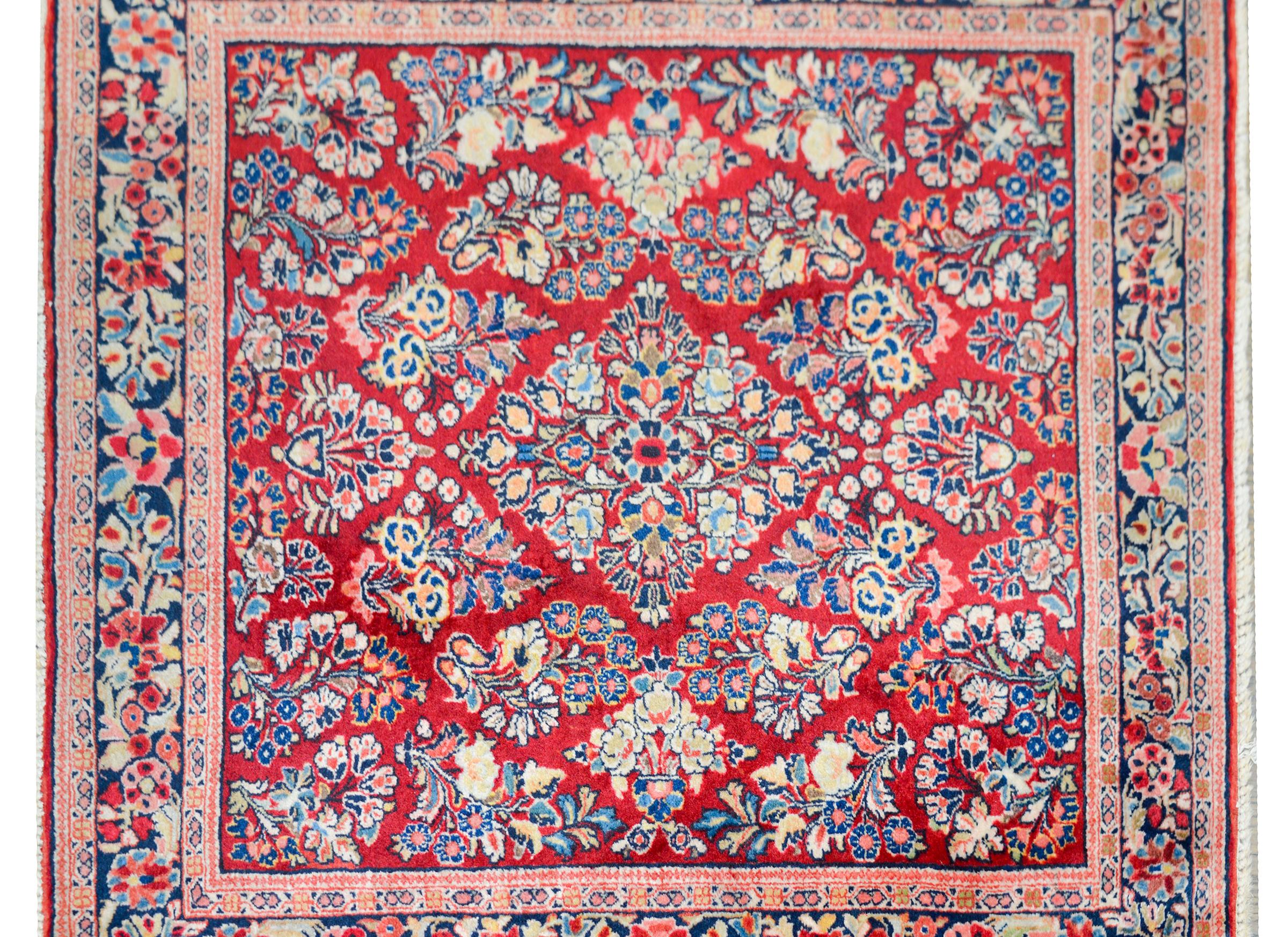 A wonderful early 20th century Persian Sarouk rug of rare square shape with an all-over mirrored floral cluster pattern woven in pink, light and dark indigo, cream, and brown, against a cranberry field, and surrounded by a complex floral border.
