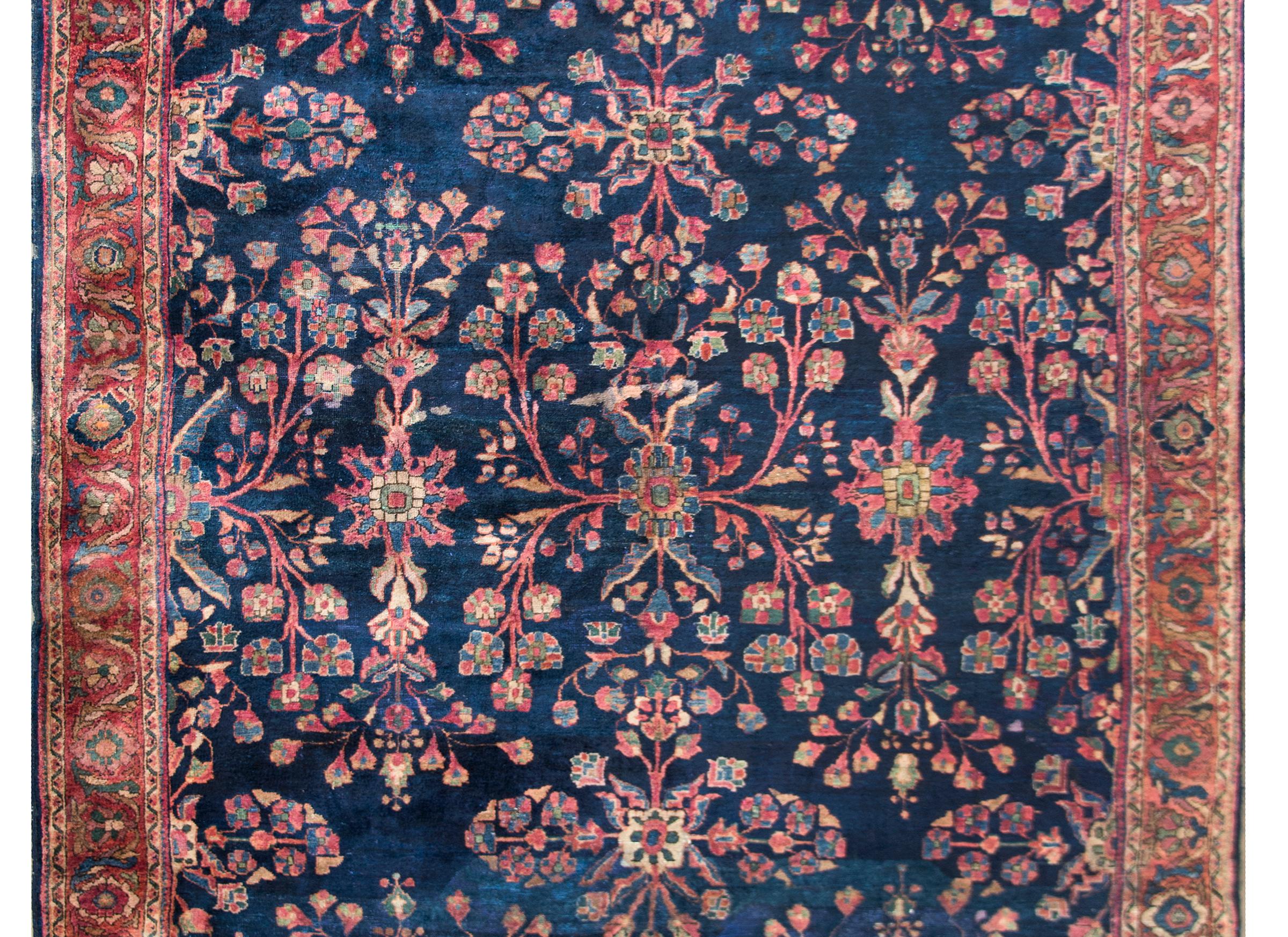 A beautiful early 20th century Persian Sarouk rug with a large-scale mirrored floral pattern woven in pinks, corals, indigos, and creams, and set against a dark indigo background. The border is sweet with a petite floral and leaf pattern, flanked by