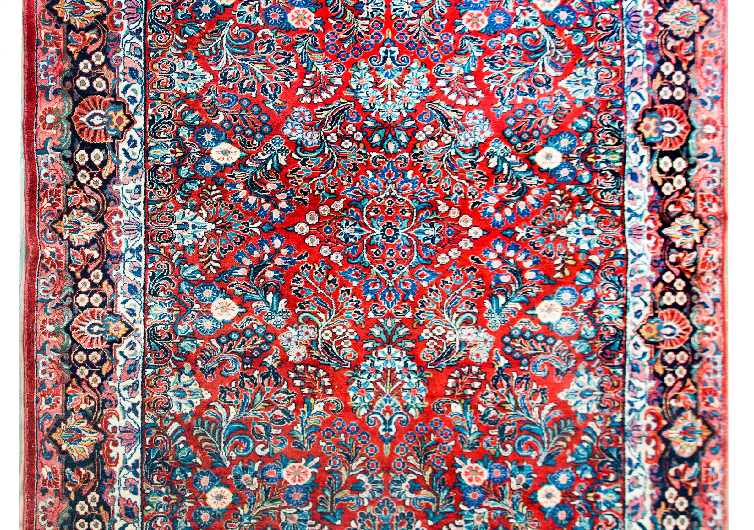 A beautiful early 20th century Persian Sarouk rug with a large-scale mirrored floral pattern containing myriad floral clusters all woven in light and dark indigo, pink, white, and yellow set against a brilliant crimson background, and surrounded by