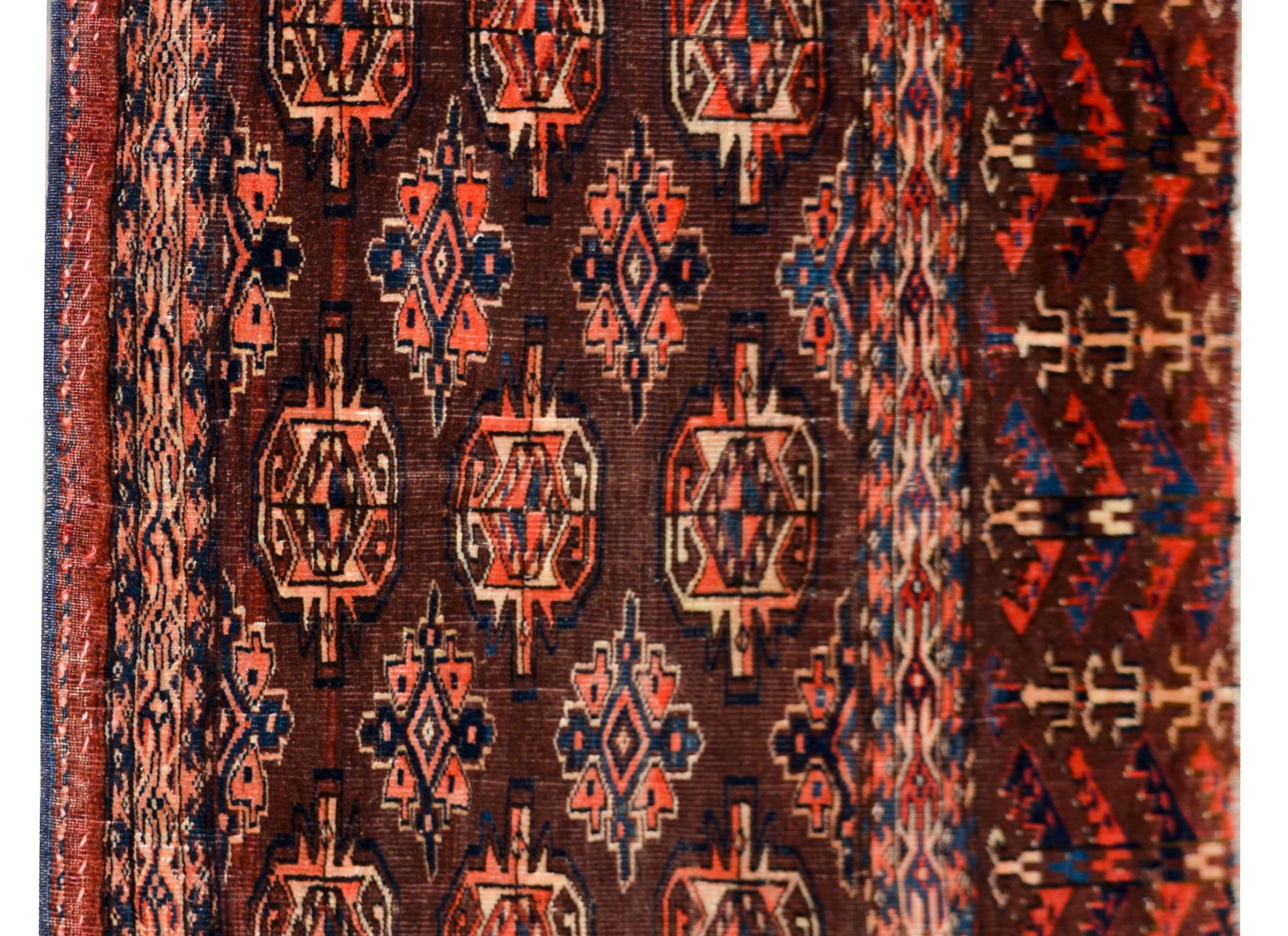 A wonderful early 20th century Persian Teke rug with an asymmetrical geometric pattern with several stylized flowers woven in crimson, indigo, and cream colored wool against a brown background, and surrounded by a border with even more geometric