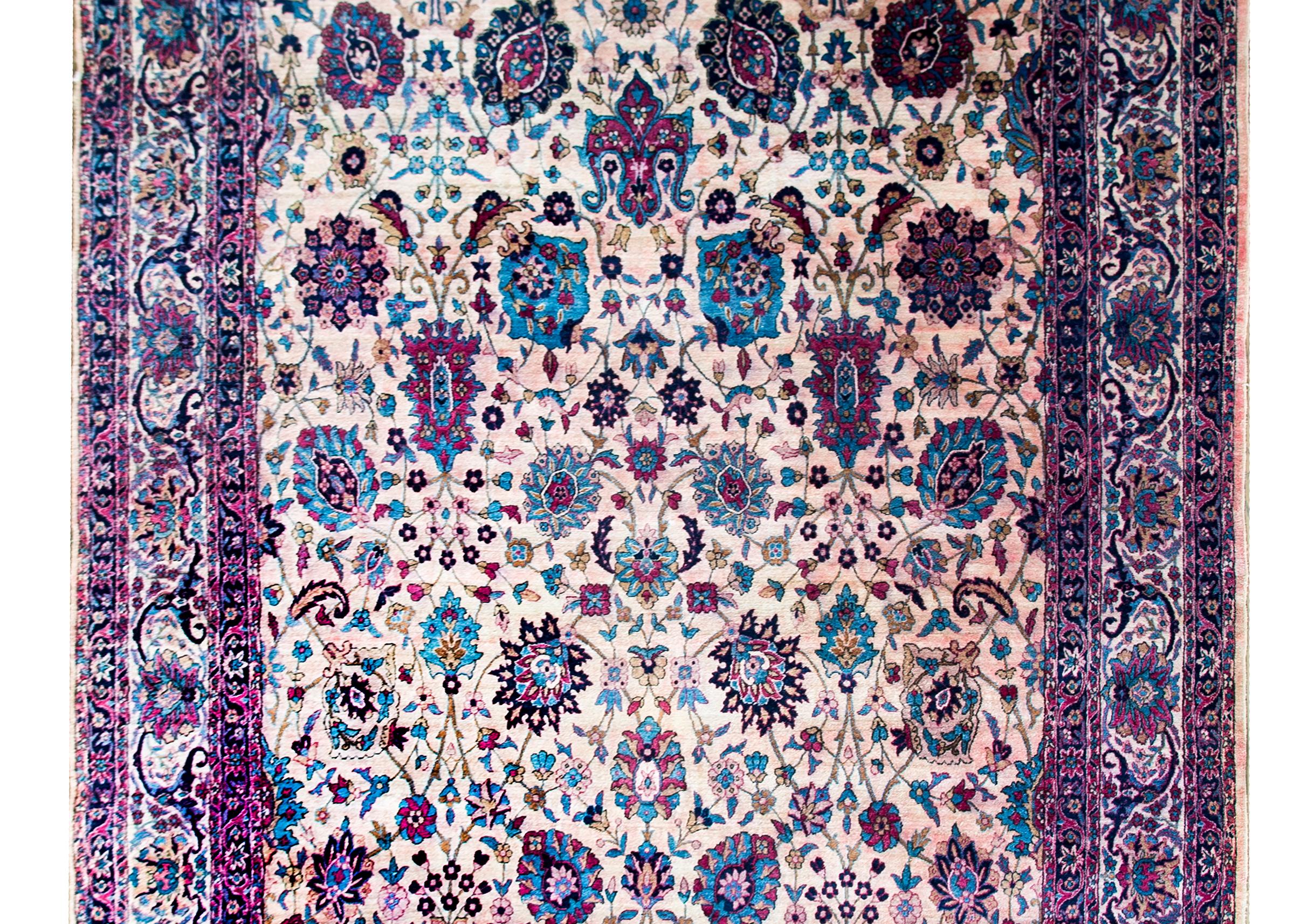 A wonderful early 20th century Persian Yazd rug with an all-over mirrored floral and scrolling vine pattern woven in light and dark indigo, cranberry, light pink, gold, and white wool set against a cream colored background.  The border is complex