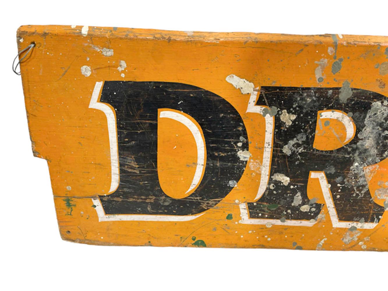 American folk art trade sign from an apothecary or pharmacy with the word 'DRUGS' in black paint with white shadow lines on an orange ground. Painted on a single pine plank with notched ends.