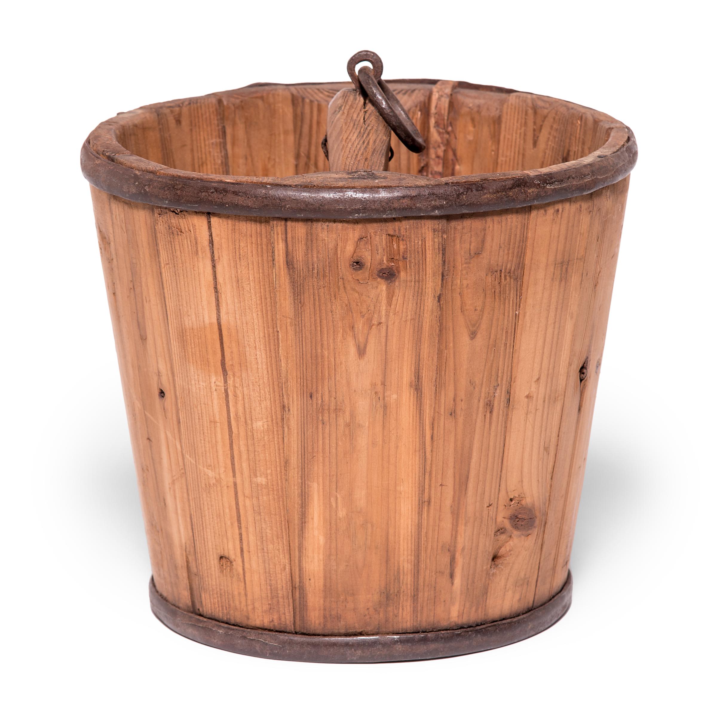 Beautifully aged with warm coloring, this Qing-dynasty bucket draws attention to the ingenuity of its manufacture, consisting of staves of pine bound with iron rings. A small ring attached to the handle suggests the bucket was once used to lift