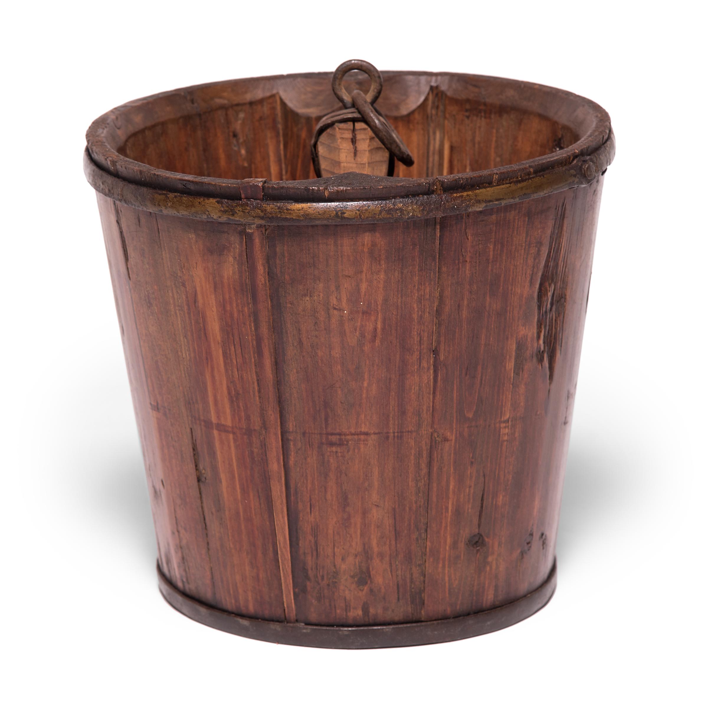 Beautifully aged with warm coloring, this Qing dynasty bucket draws attention to the ingenuity of its manufacture, consisting of staves of pine bound with iron rings. A small ring attached to the handle suggests the bucket was once used to lift